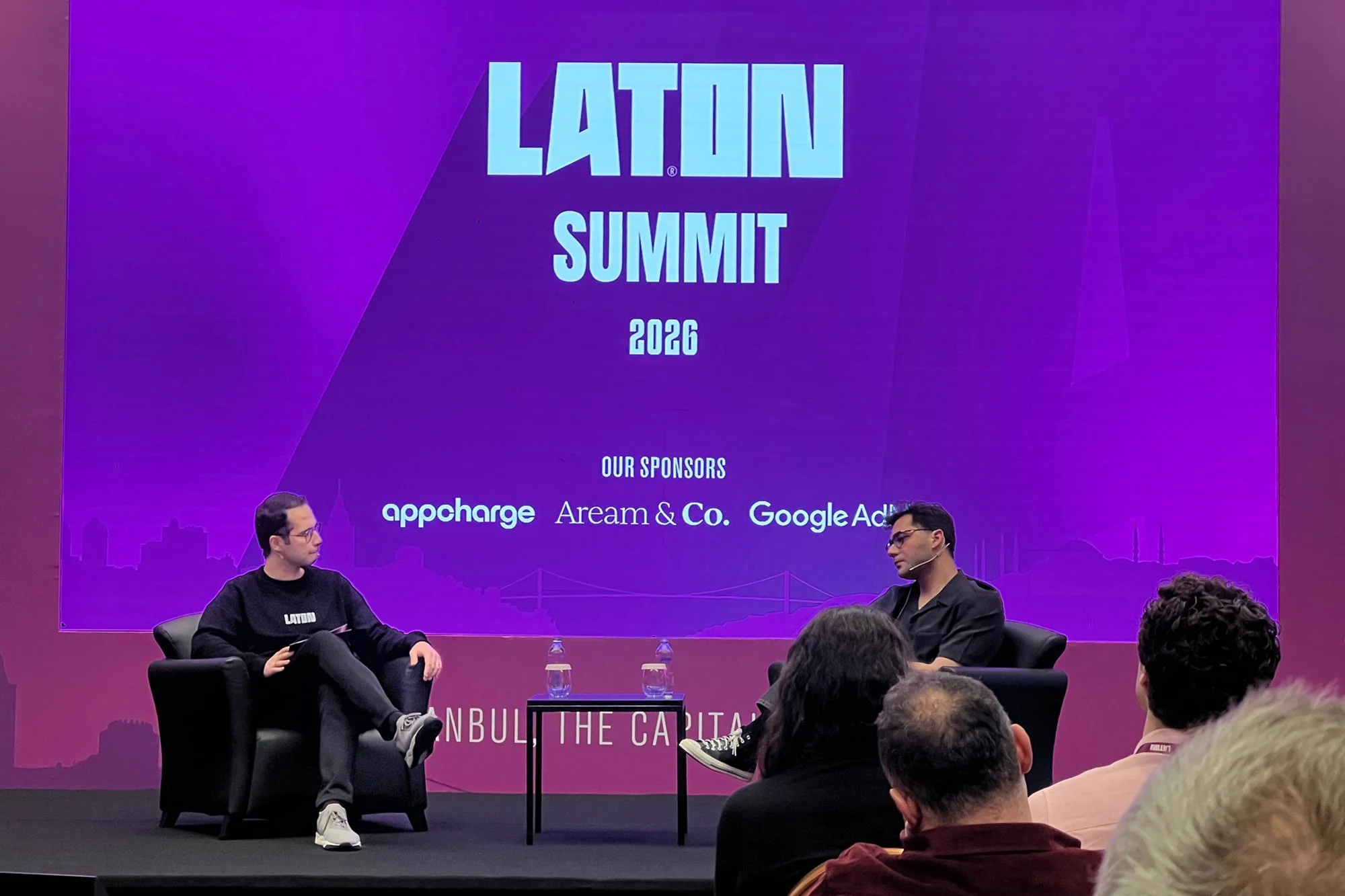 At a LATIN Summit 2026 event, two men are seated on a stage with a large purple and blue backdrop displaying the event name and sponsors, including AppChrage, Aream & Co., and Google Ads, with an audience watching.