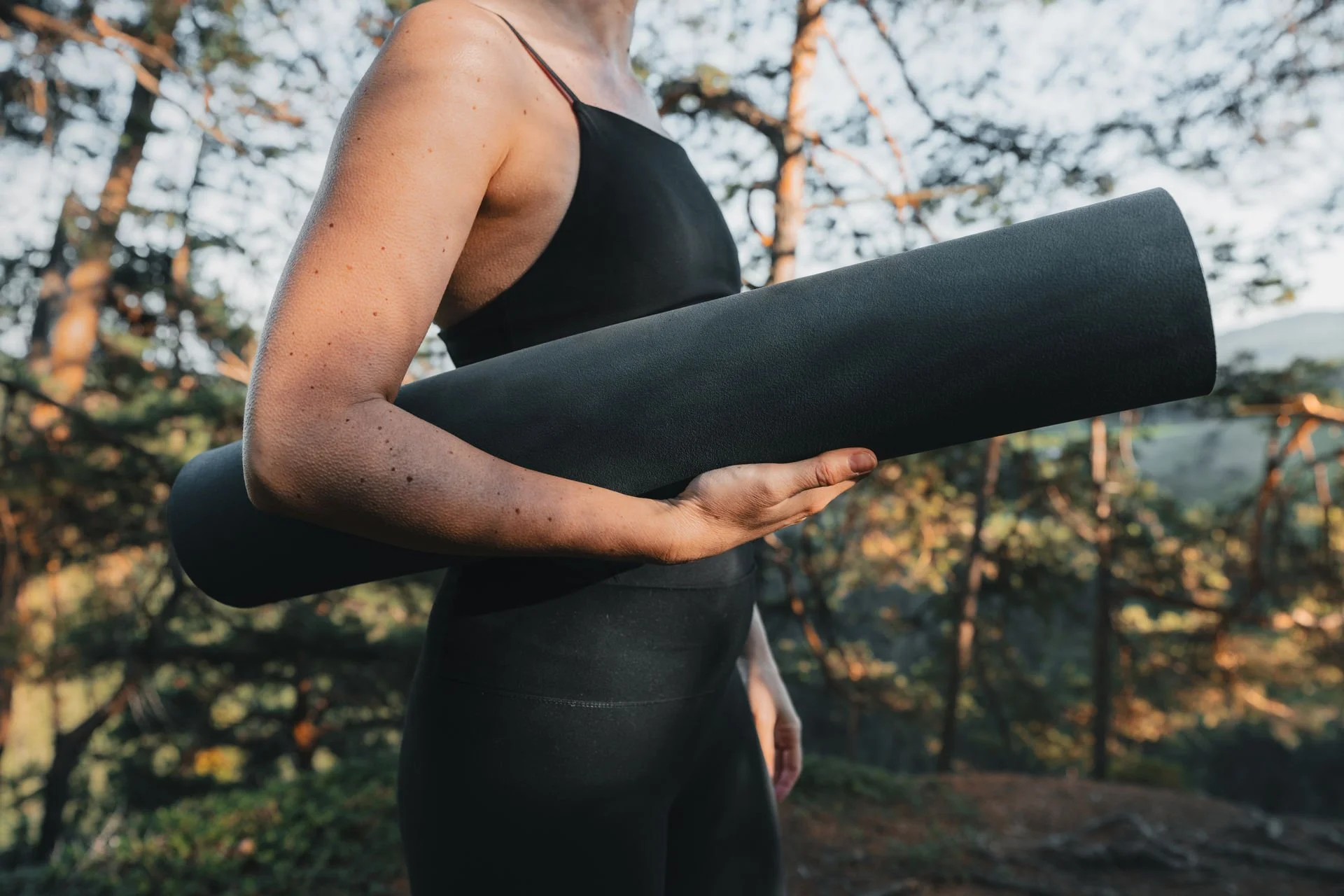 Frau trägt schwarze Yogahose und Top, hält eine schwarze Yogamatte im Freien bei Sonnenuntergang