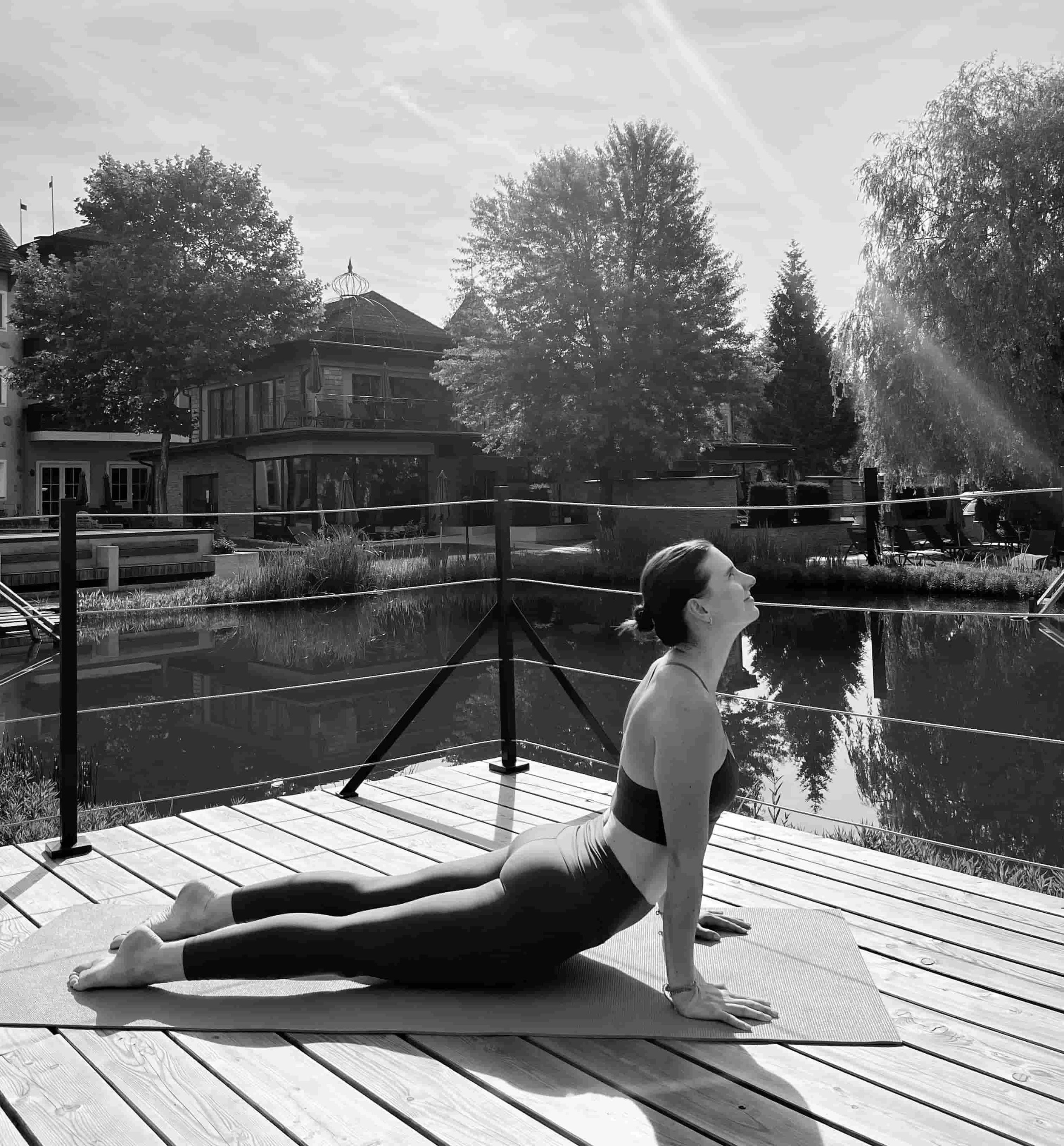 Eine Frau macht Yoga auf einer Holzplattform am Wasser, die Pose ist der Cobra. Die Szene ist im Freien mit Bäumen und Gebäuden im Hintergrund, die Sonne scheint.