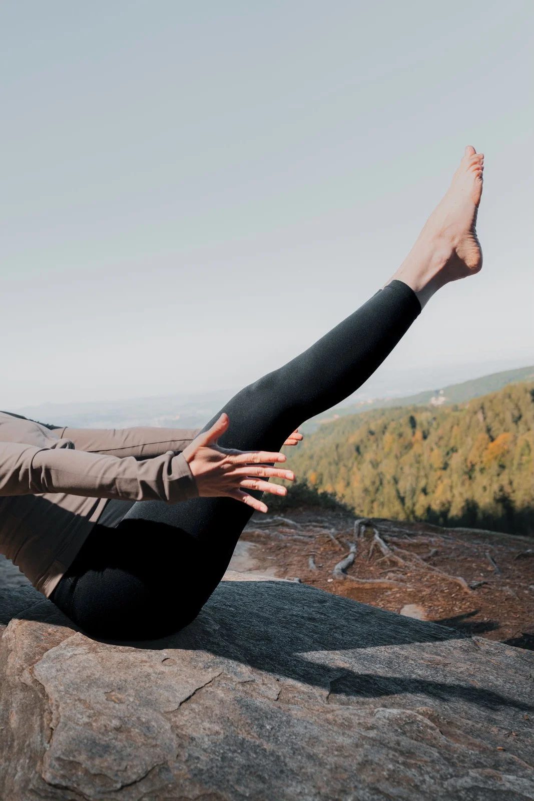 Person macht eine Yoga-Übung auf einem Felsen, mit den Beinen in die Luft gestreckt, im Freien mit Blick auf einen bewaldeten Hügel und einen klaren Himmel.