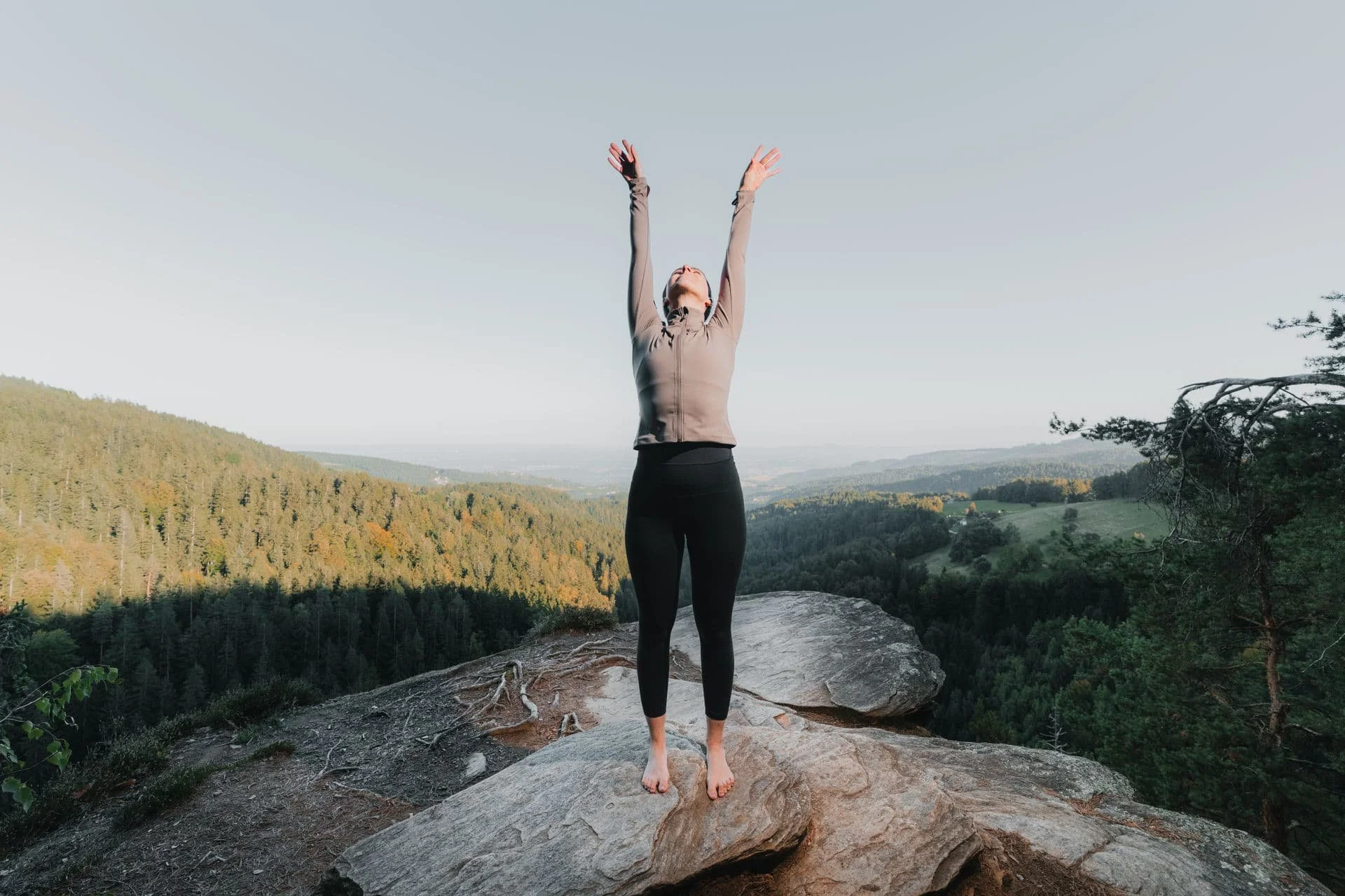 Eine Frau steht barfuß auf einem Felsen auf einem Hügel mit Blick auf einen Wald und eine offene Landschaft, hebt die Arme hoch und wirkt entspannt.