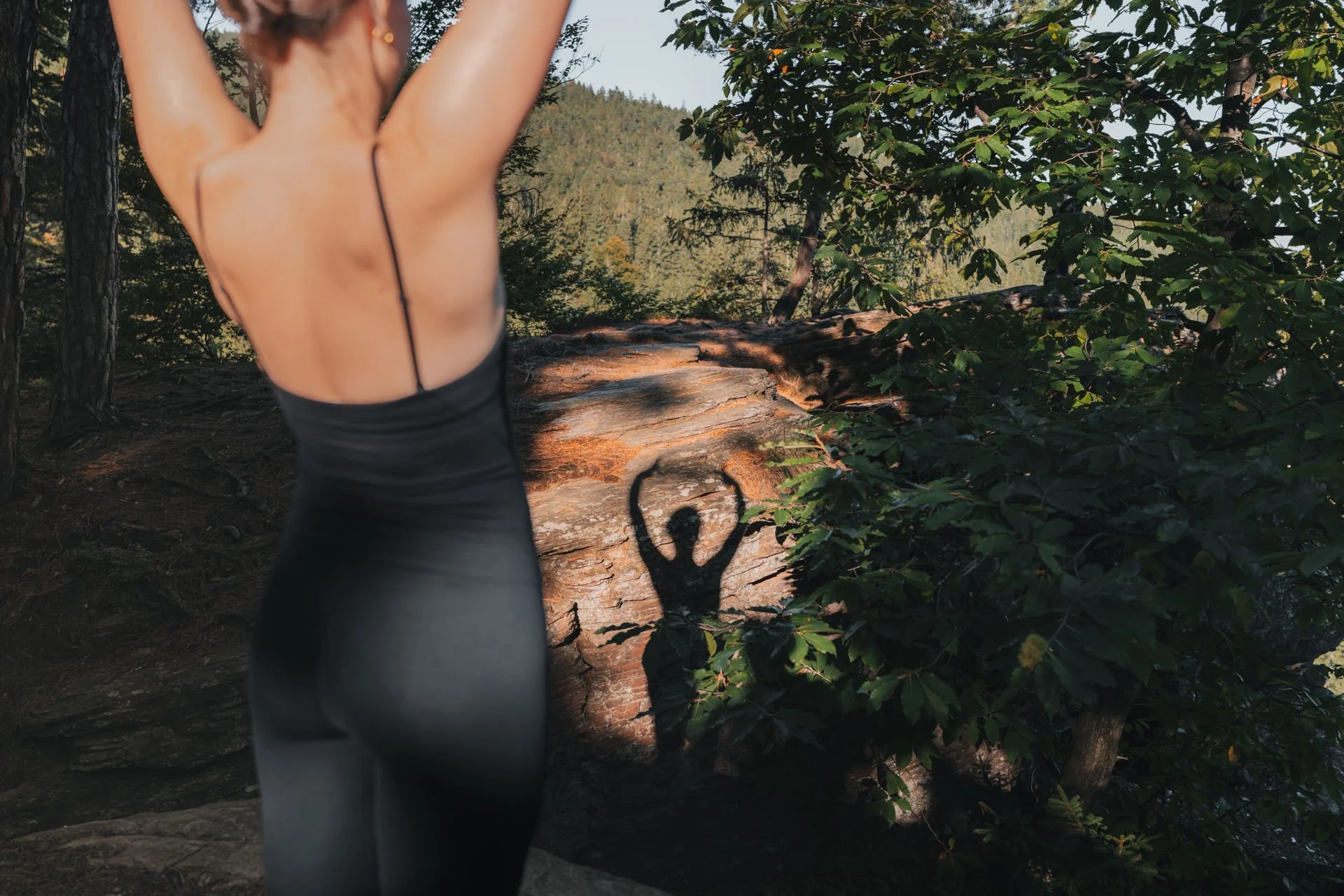 Frau beim Yoga im Freien, Schatten ihrer Arme und Hände in der Nähe eines Felsens im Wald, umgeben von Bäumen und grünen Blättern, Sonnenlicht