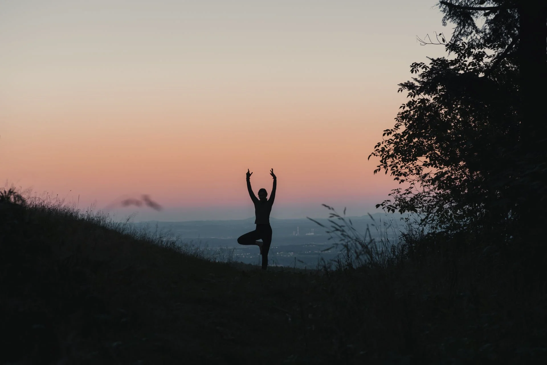 Eine Person, die im Baumkreis Yoga macht, im Sonnenuntergang auf einem Hügel, mit einem farbigen Himmel im Hintergrund.