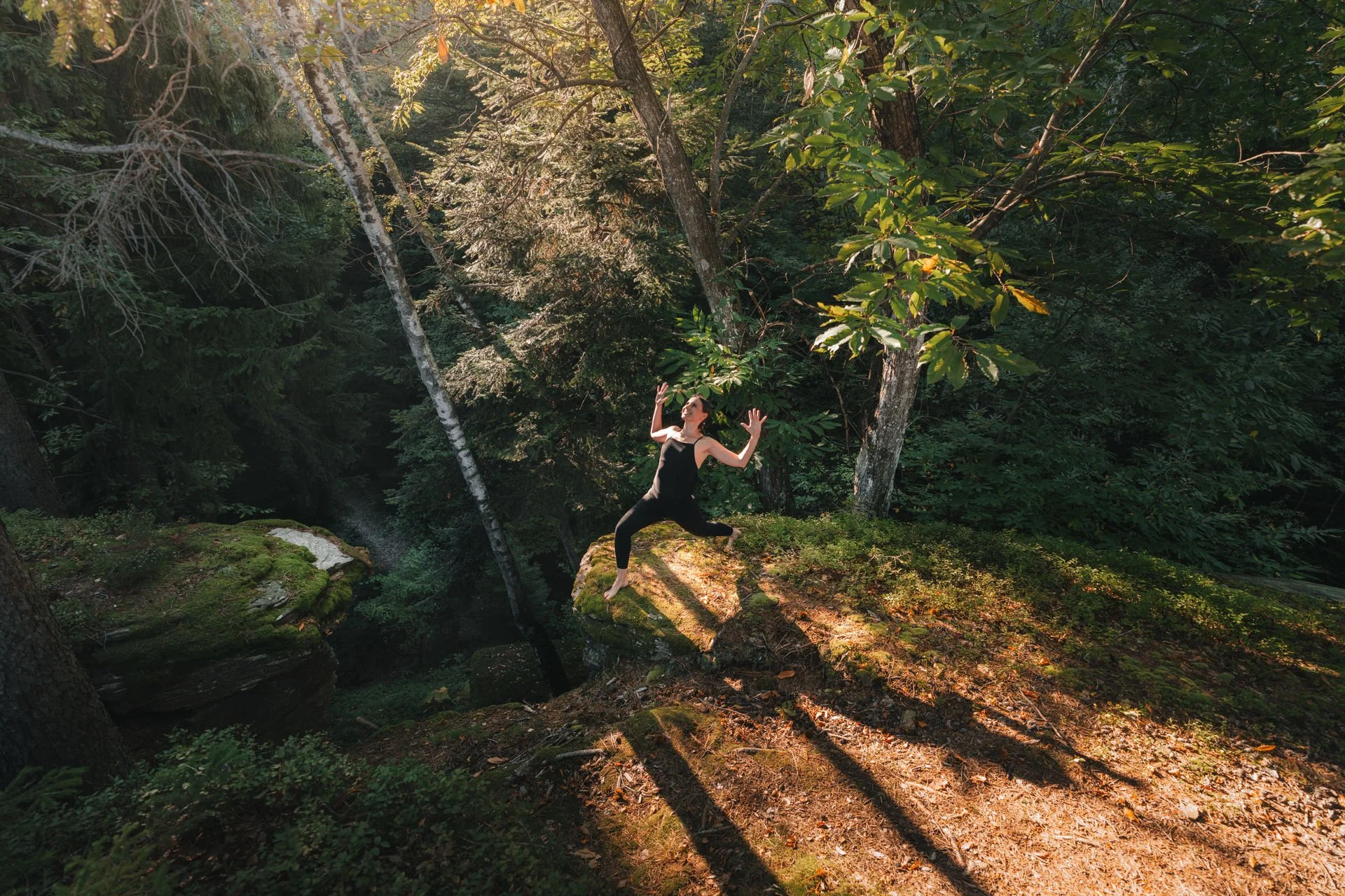 Eine Frau in schwarzer Kleidung steht auf einem moosbedeckten Felsen in einem grünen Wald, die Arme erhoben, im Sonnenlicht.
