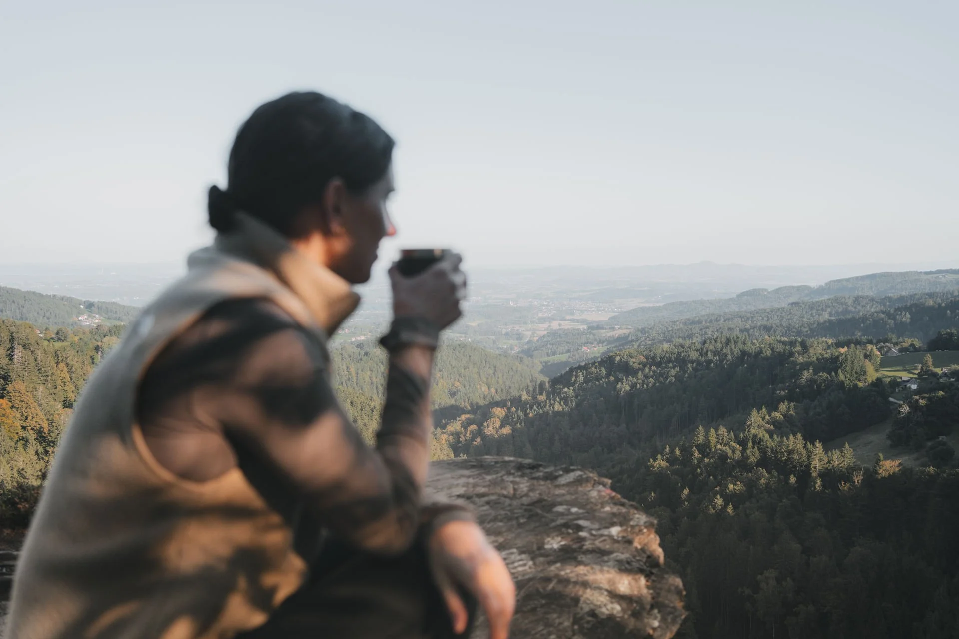 Ein Mann sitzt auf einem Felsen und trinkt aus einer Tasse, während er die Aussicht auf die Hügel und Wälder im Hintergrund genießt.