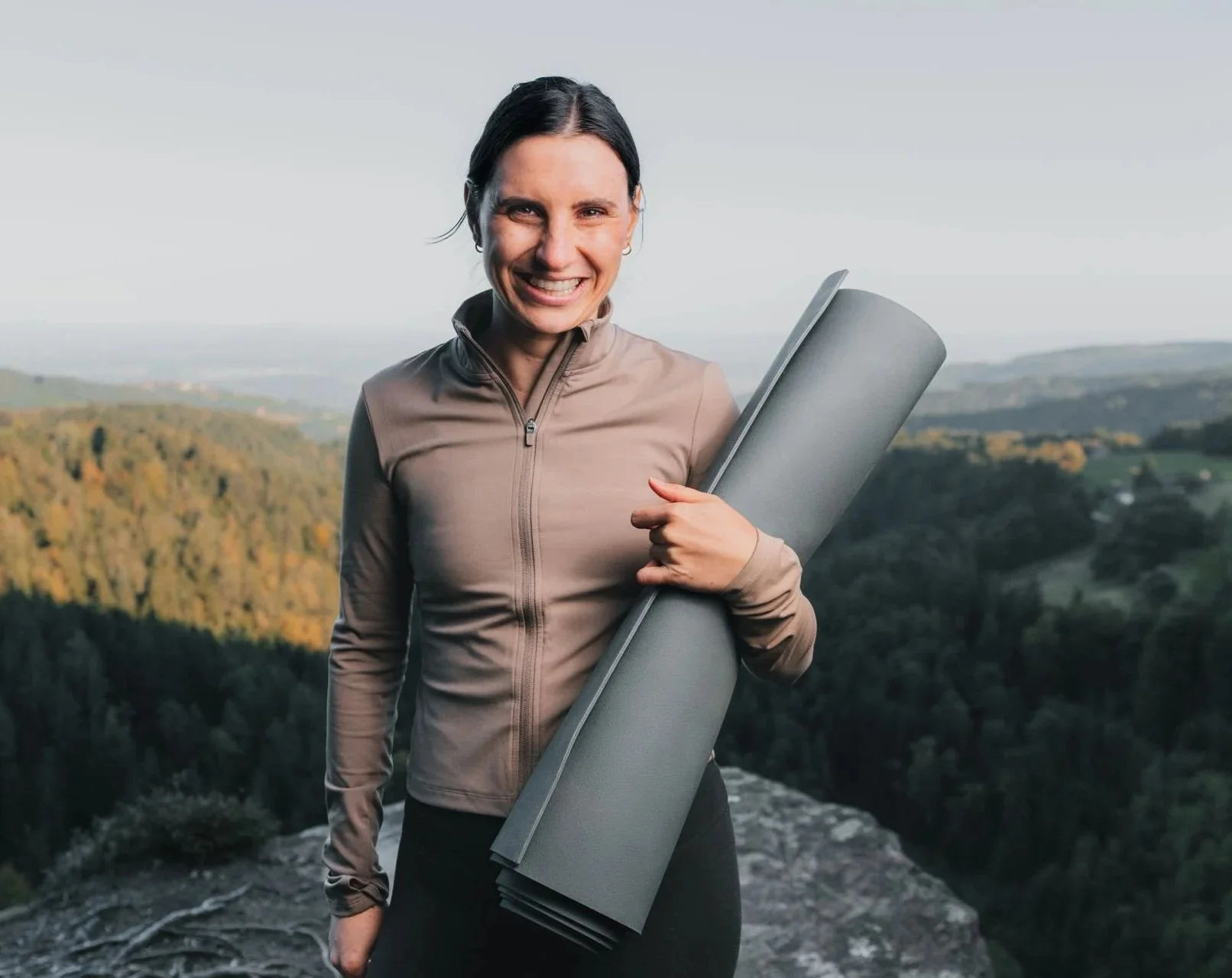 Frau steht auf einem Berggipfel, hält eine Yogamatte und lächelt in die Kamera, im Hintergrund eine grüne Landschaft mit Bäumen und Hügeln.