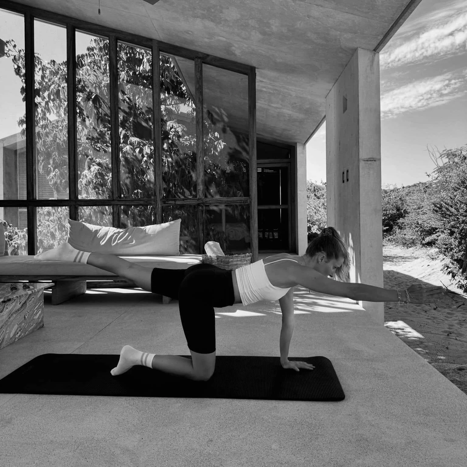 Frau macht Yoga auf einer Matte in einem modernen, offenen Raum mit großen Fenstern, die eine Gartenlandschaft im Hintergrund zeigen.