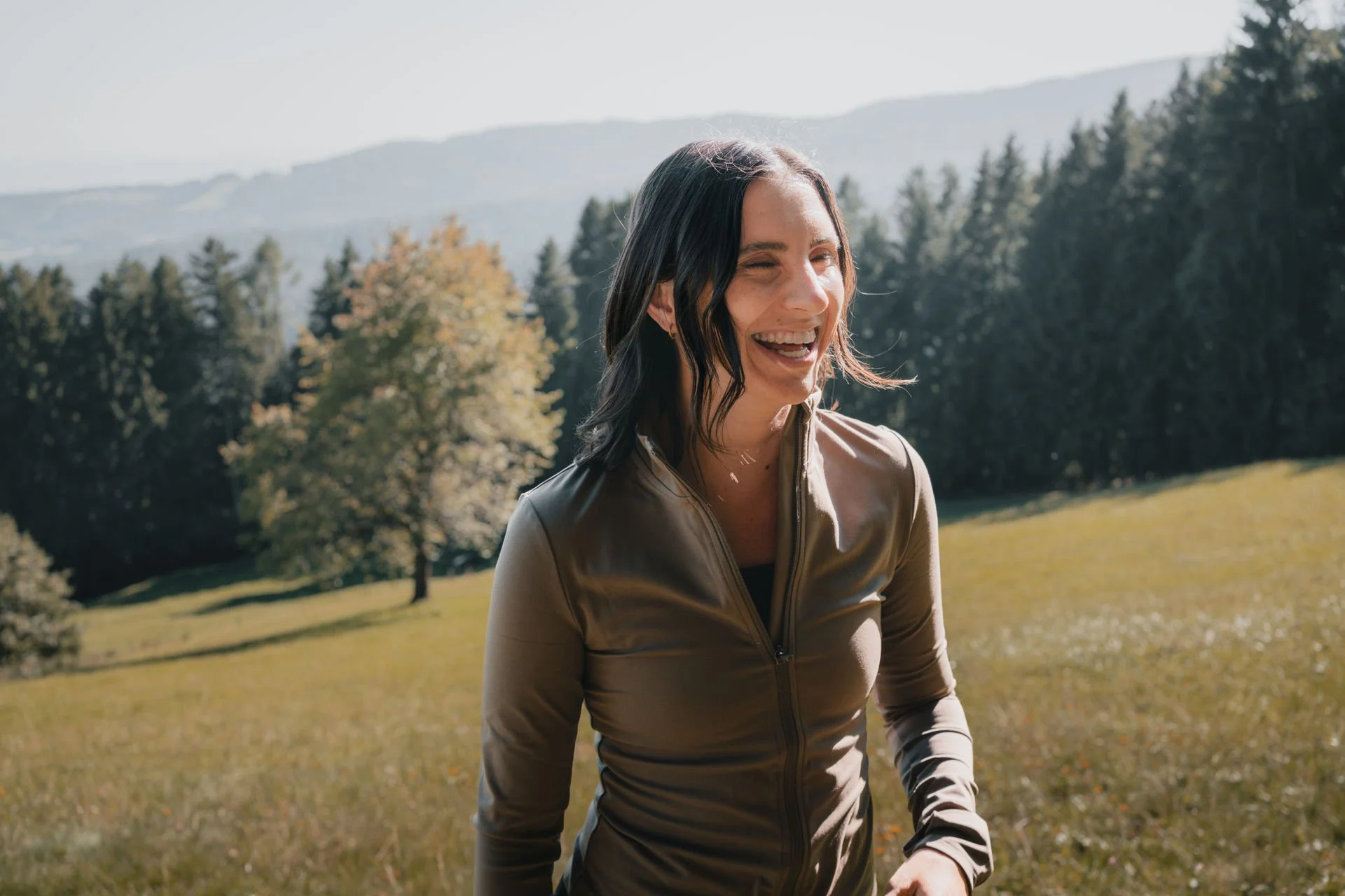 Eine lachende Frau mit dunklen, gewellten Haaren, trägt eine beige Jacke, im Freien in einer grünen, bewaldeten Landschaft.