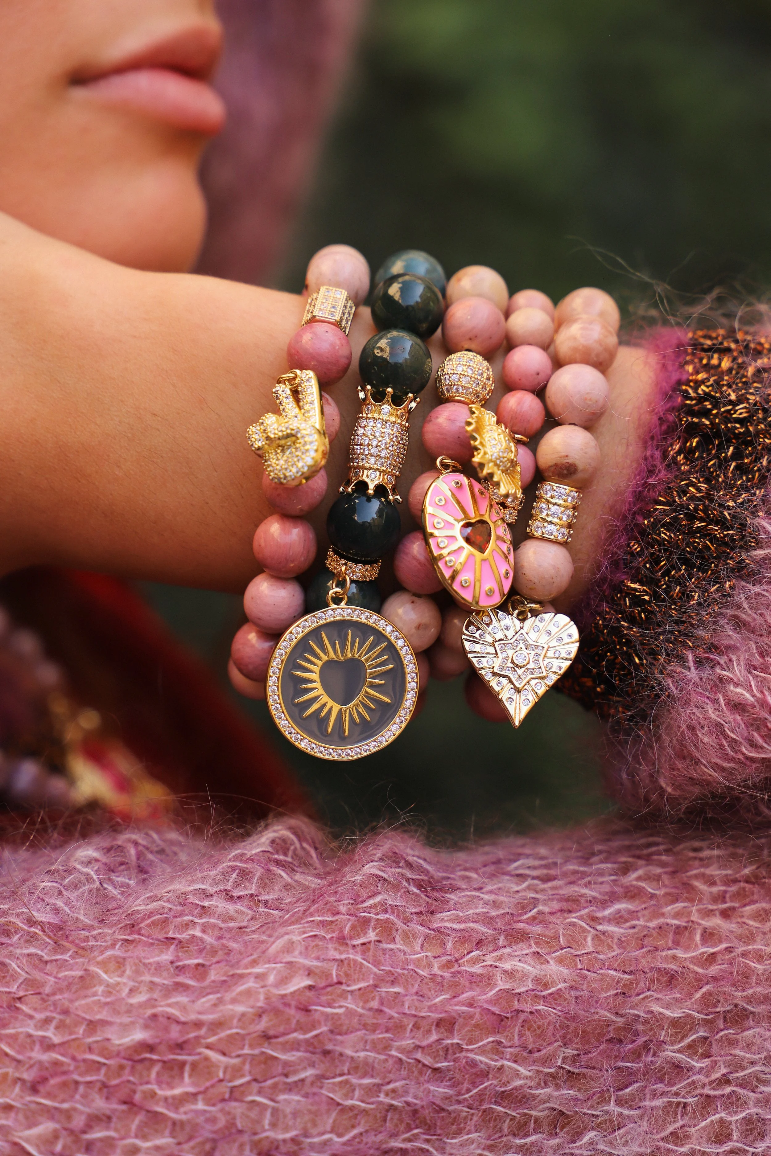 Une main féminine portant plusieurs bracelets colorés avec des pendentifs en or et en pierre, notamment un pendentif avec un soleil, un cœur orné de pierres, et d'autres pendentifs en forme de cœur, d'anneau, et de personnages.