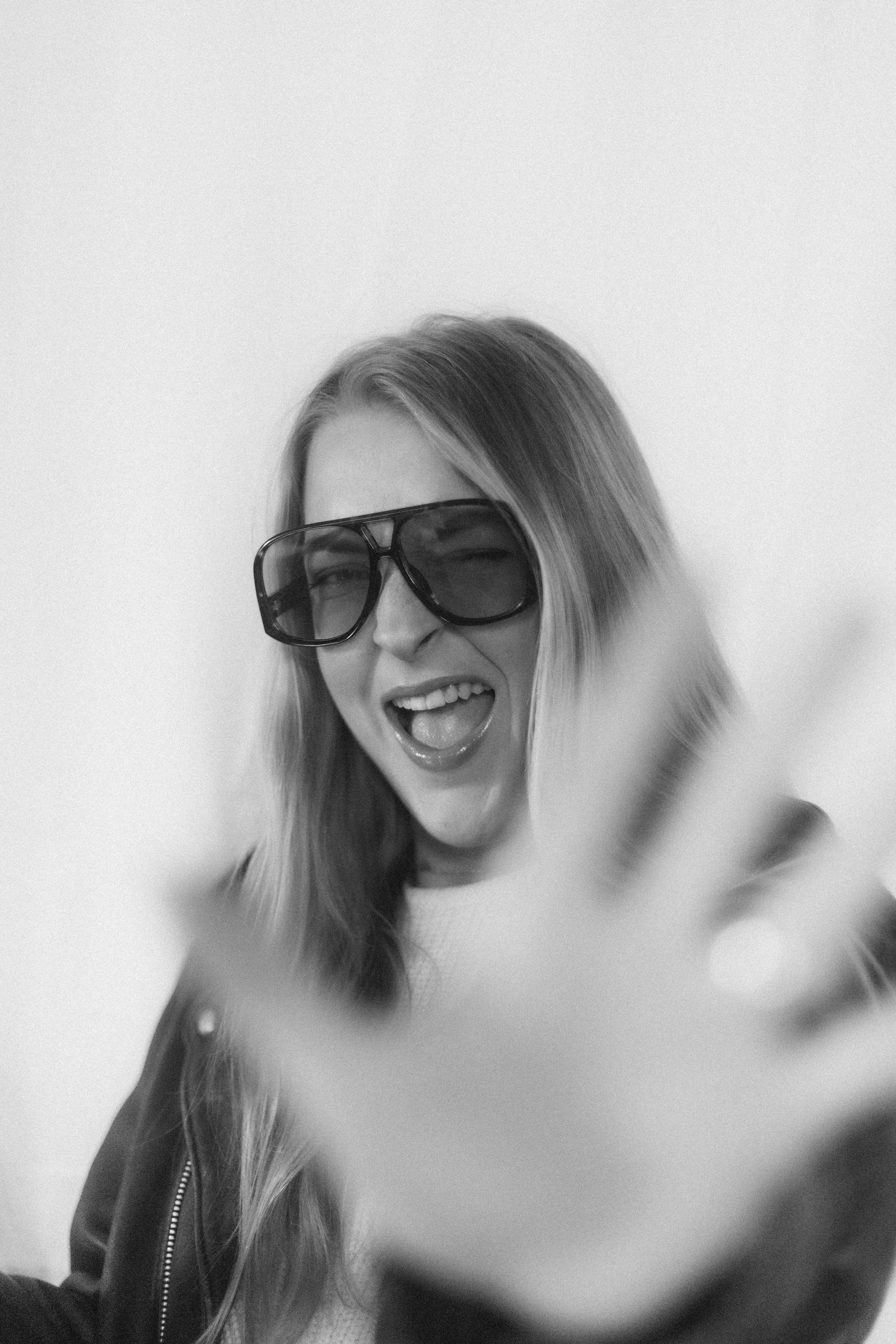 Jeune femme souriante portant des lunettes de soleil sombres, faisant un clin d'œil, avec une main en premier plan et un fond blanc.