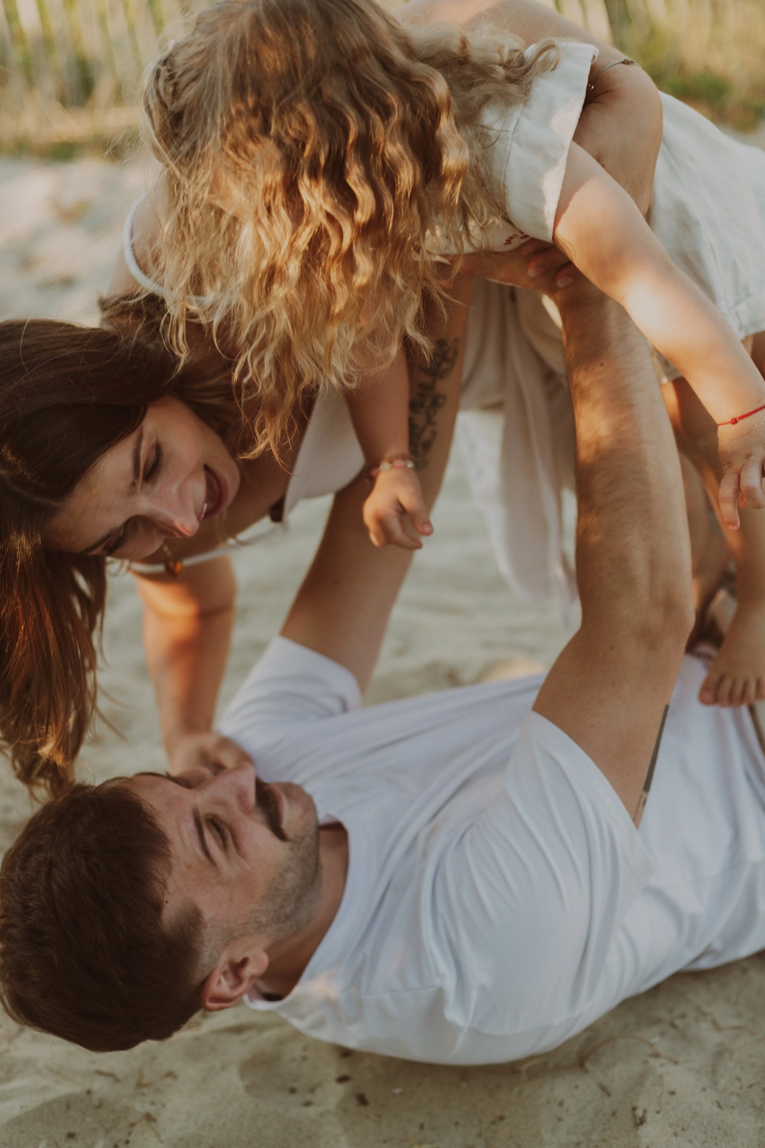 Une famille de quatre personnes jouant ensemble sur la plage, avec un père allongé sur le sol, une mère aidant une fille avec des cheveux roux, le tout sur du sable, lors d'une journée ensoleillée.