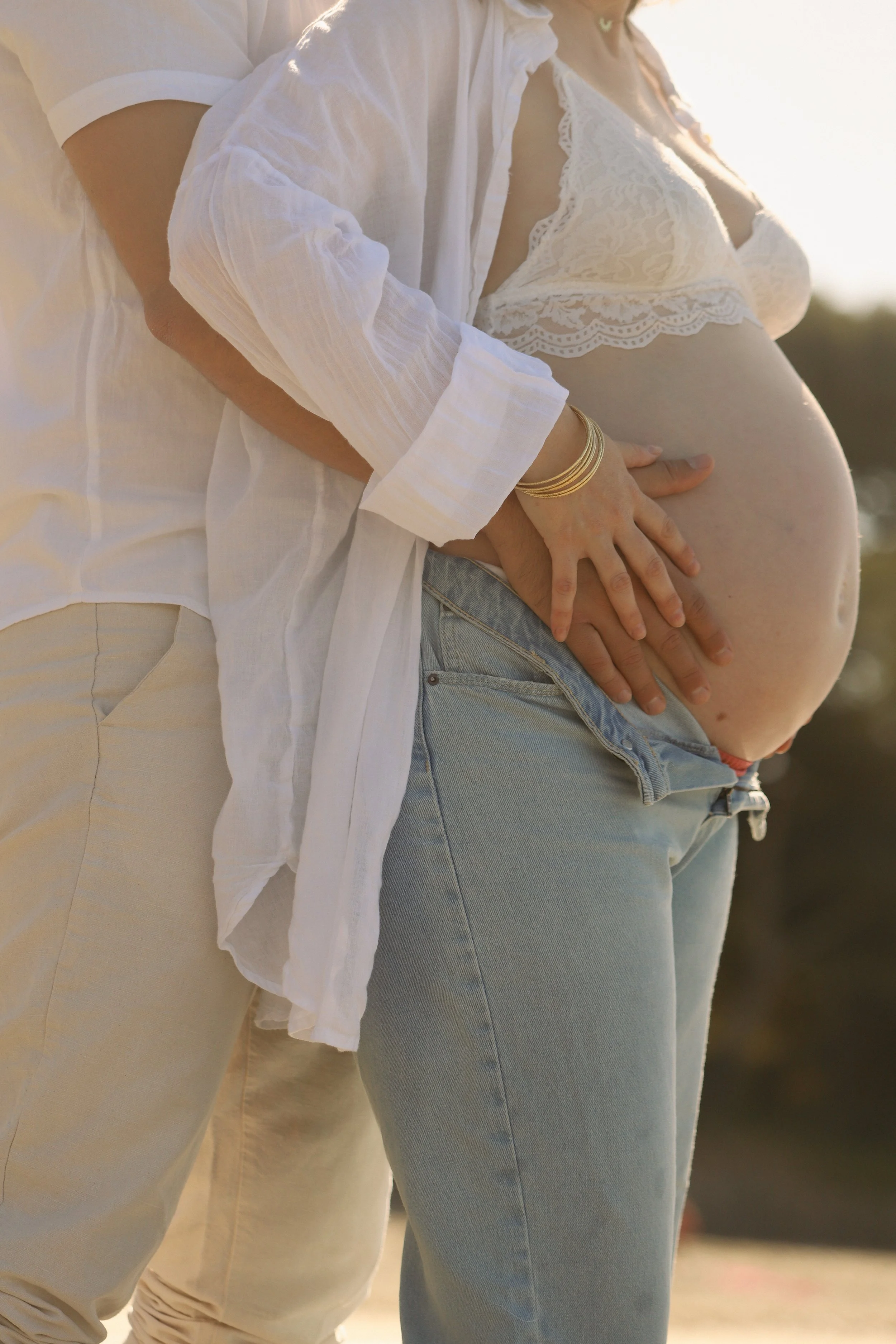 Une femme enceinte portant un haut en dentelle blanc et un homme derrière elle, ils tiennent son ventre de mains jointes, en plein air.