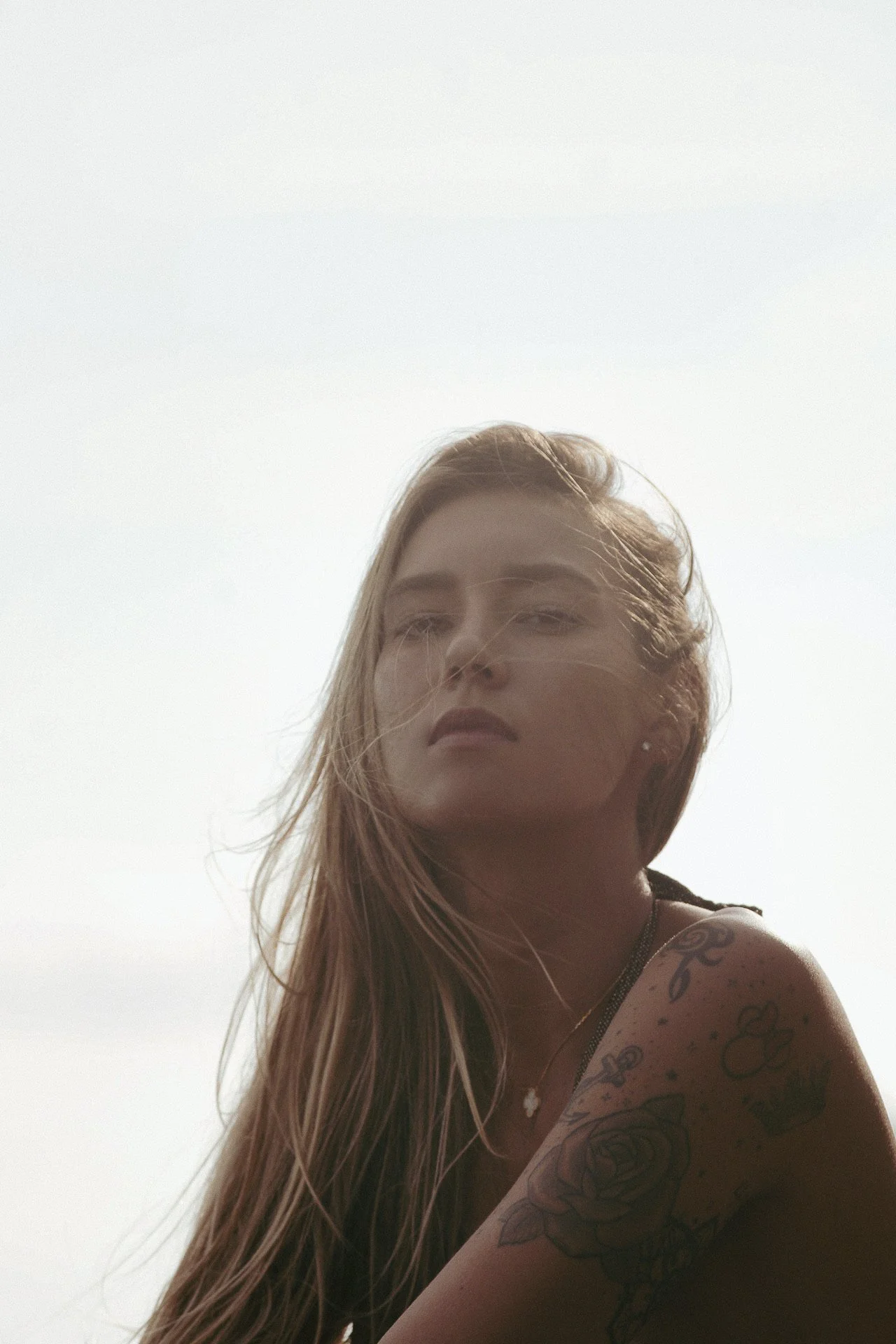 Jeune femme avec tatouages sur l'épaule, cheveux longs et bouclés, posant en extérieur avec un ciel clair en arrière-plan.