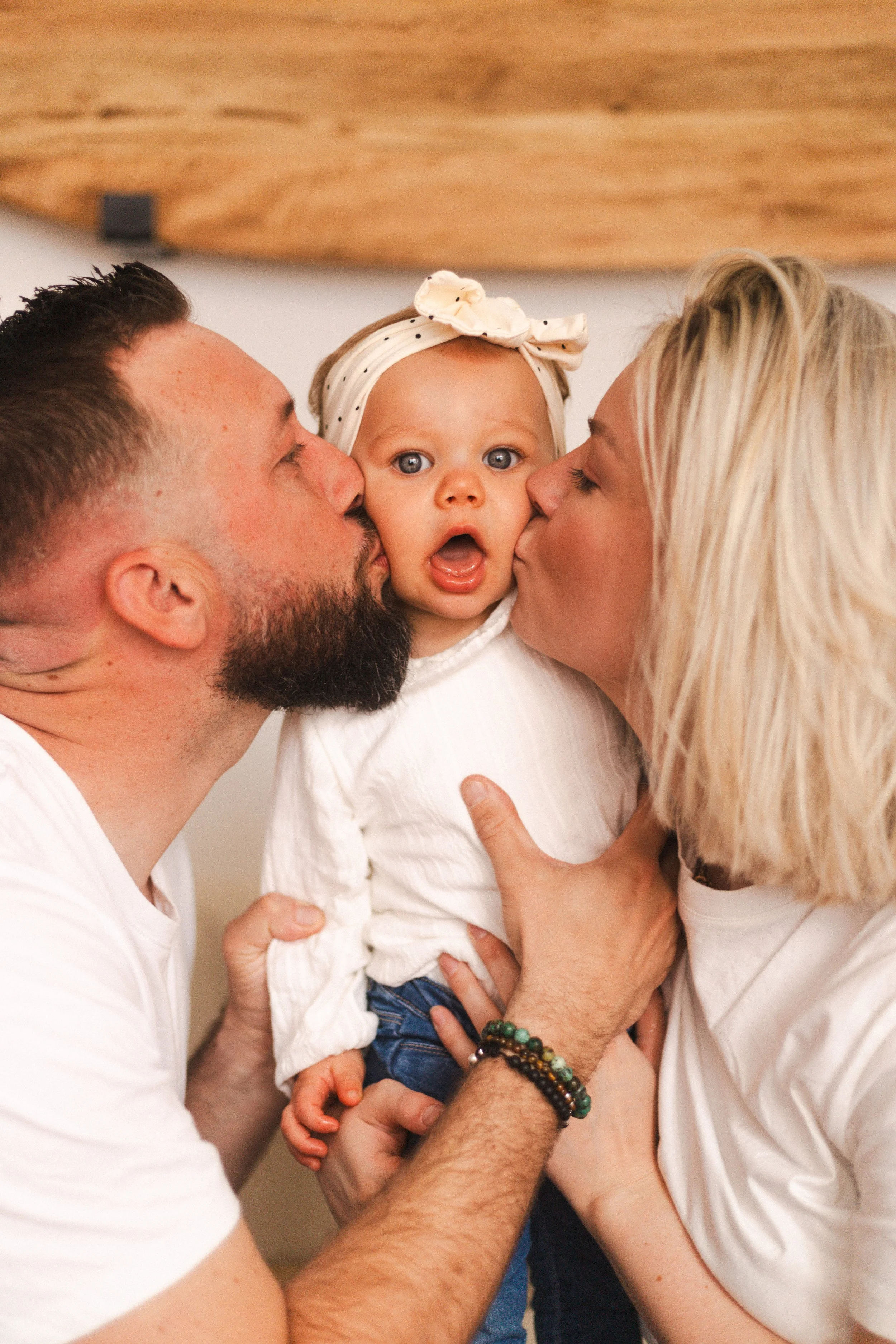 Un enfant est embrassé par un homme et une femme, tous deux lui donnent un baiser sur chaque joue. La fille a les yeux grands ouverts et porte un bandeau avec un nœud blanc.