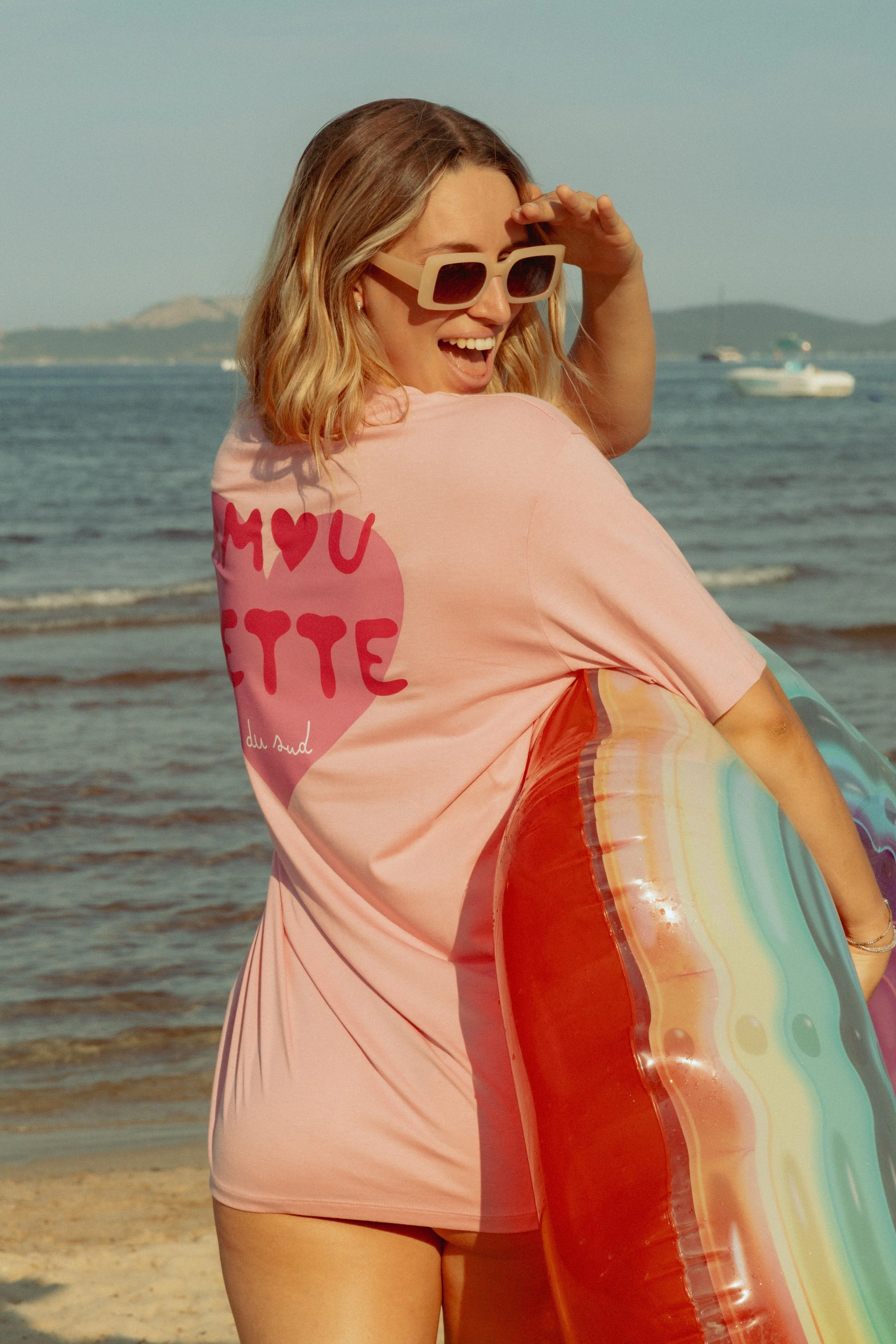 Femme souriante à la plage avec un ballon gonflable en forme de gâteau arc-en-ciel, portant des lunettes de soleil et un t-shirt rose avec un message en français.