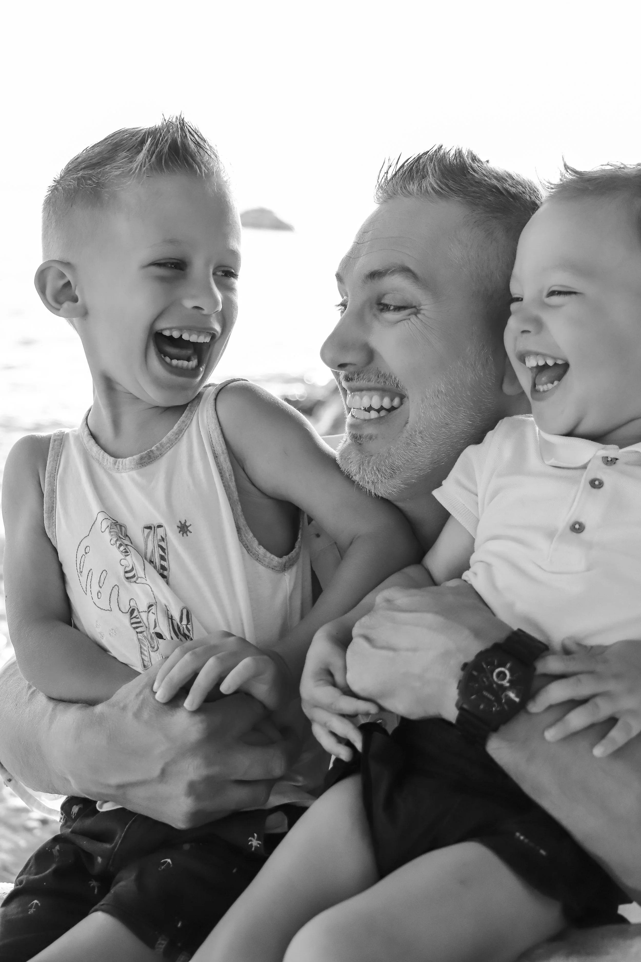 Un homme et deux jeunes garçons souriants, partageant un moment joyeux au bord de la mer.