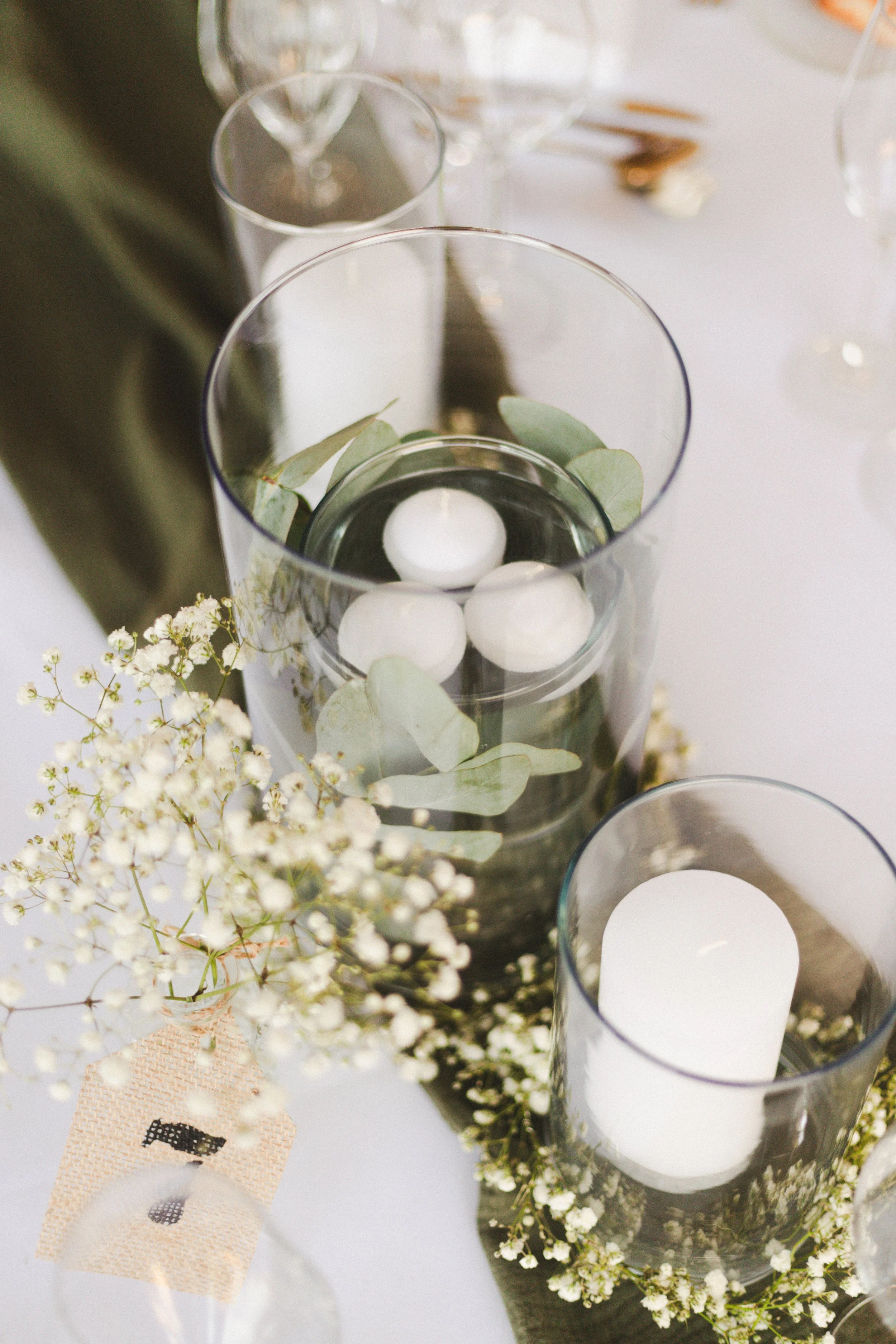Décoration de table avec des bougies dans des vases en verre, entourées de feuilles vertes et de fleurs blanches.