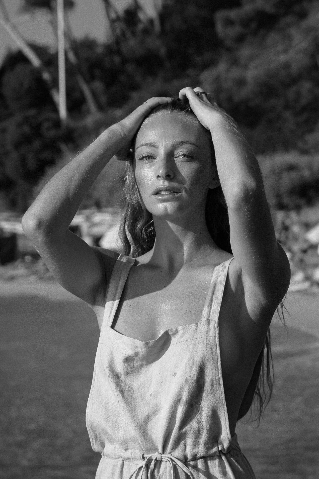 Une femme sur la plage, yeux mi-clos, portant une robe sans manches, cheveux longs, mains dans ses cheveux, style portrait en noir et blanc.