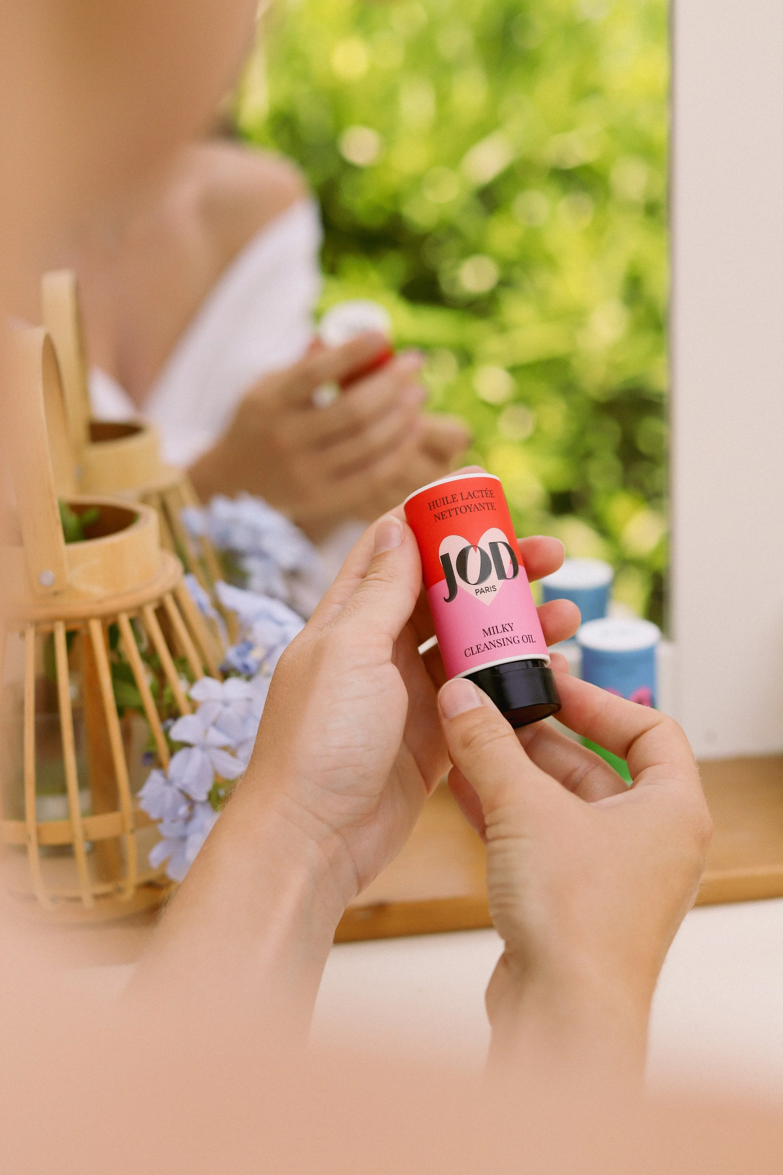 Une main tient un flacon de produit nettoyant pour le visage devant un miroir, avec une personne se nettoyant le visage en arrière-plan.