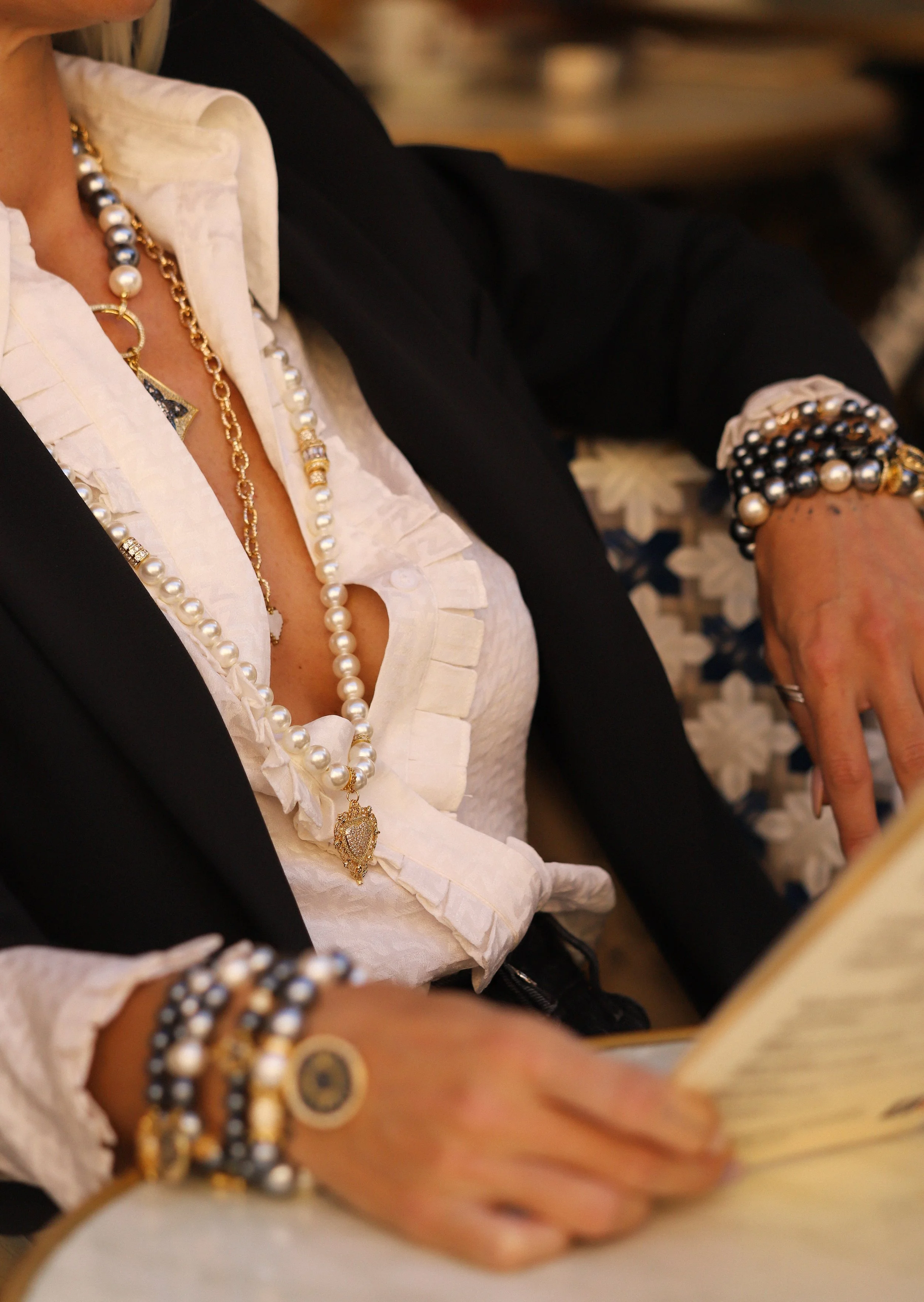 Une personne portant plusieurs bracelets, un collier de perles et un vêtement blanc avec un veston noir, assise et lisant un livre ou une carte.