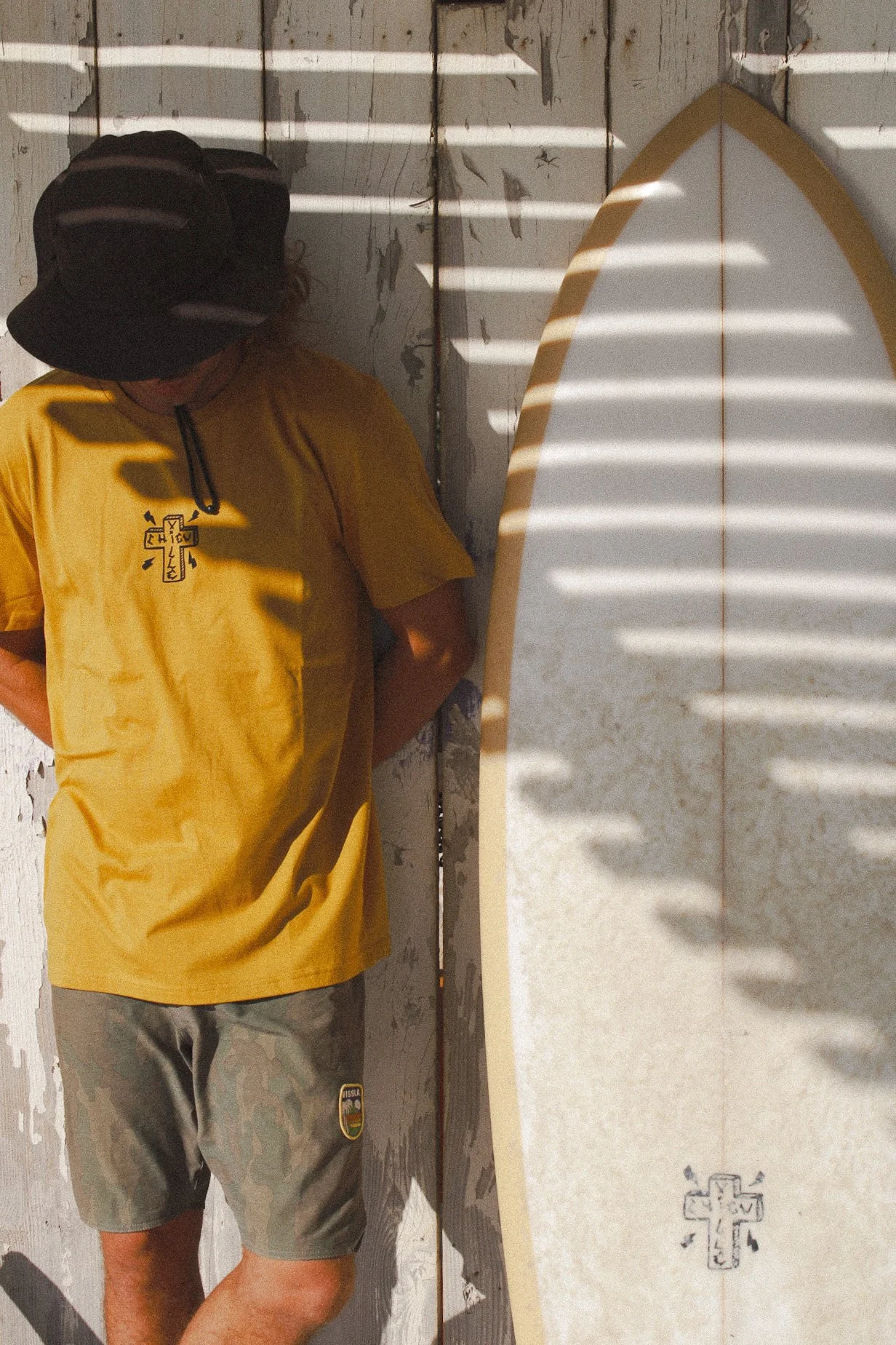 Jeune homme en t-shirt jaune et short gris, portant un chapeau noir, debout contre un mur en bois peint, avec une planche de surf à côté, et des rayures de lumière au soleil créant un motif de lignes blanches.