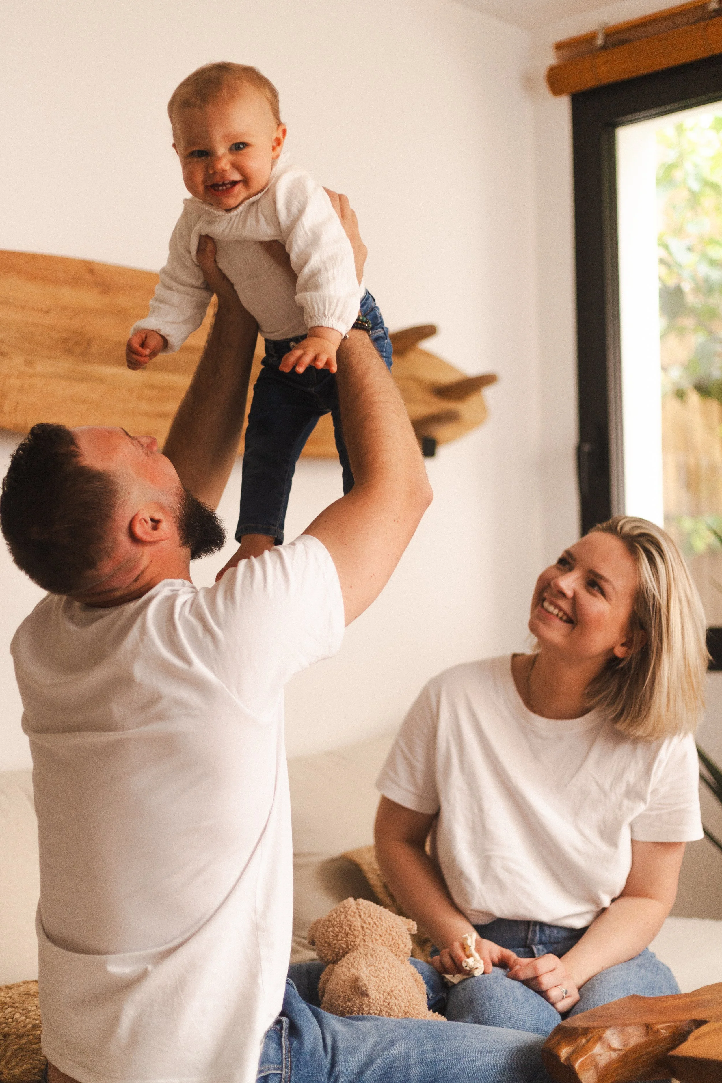 Un homme soulève un jeune enfant dans une pièce lumineuse pendant que deux femmes regardent et sourient.