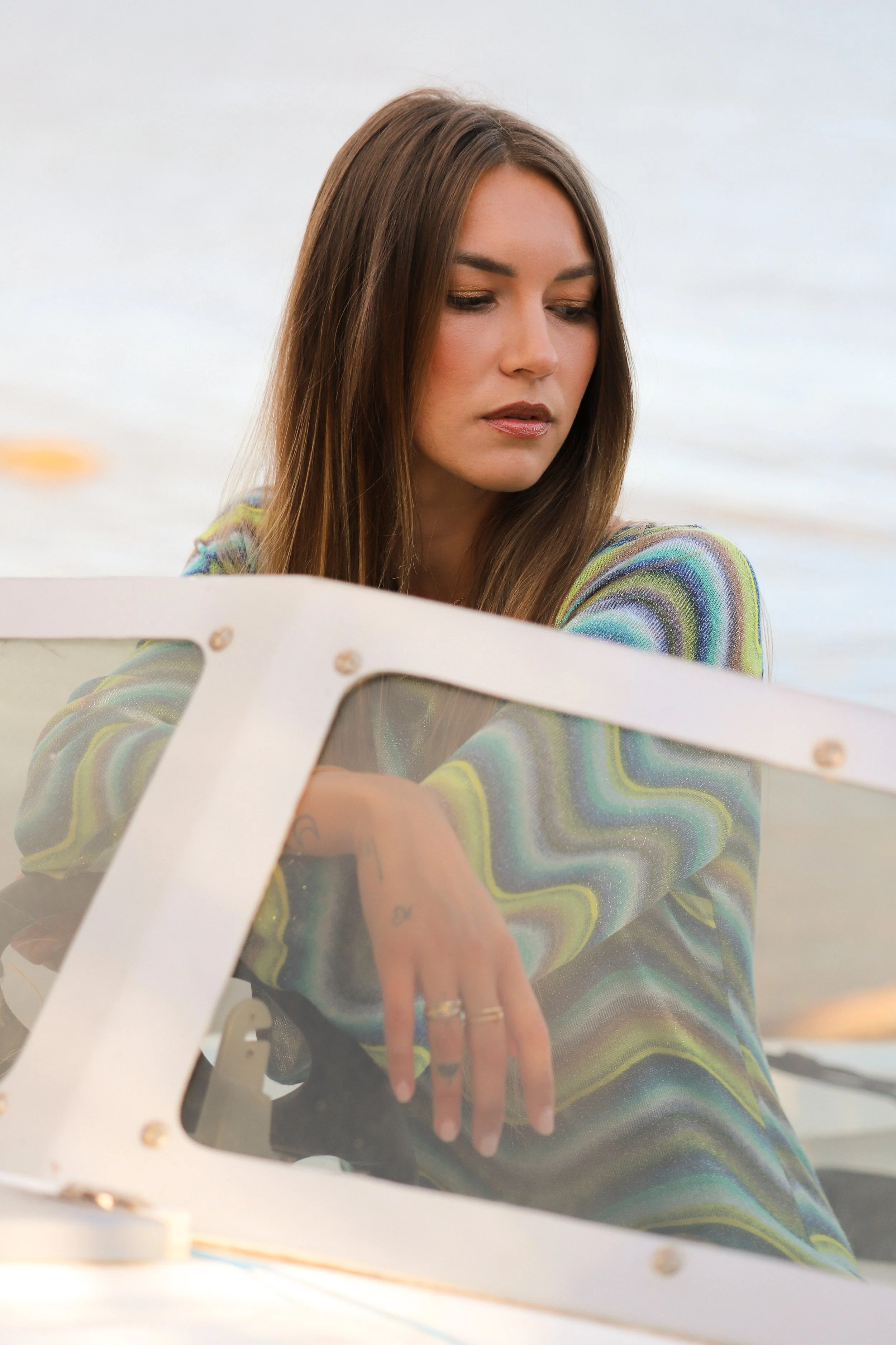 Une femme aux cheveux longs et bruns regarde à travers une fenêtre en verre, en se trouvant à l'extérieur près de l'eau.