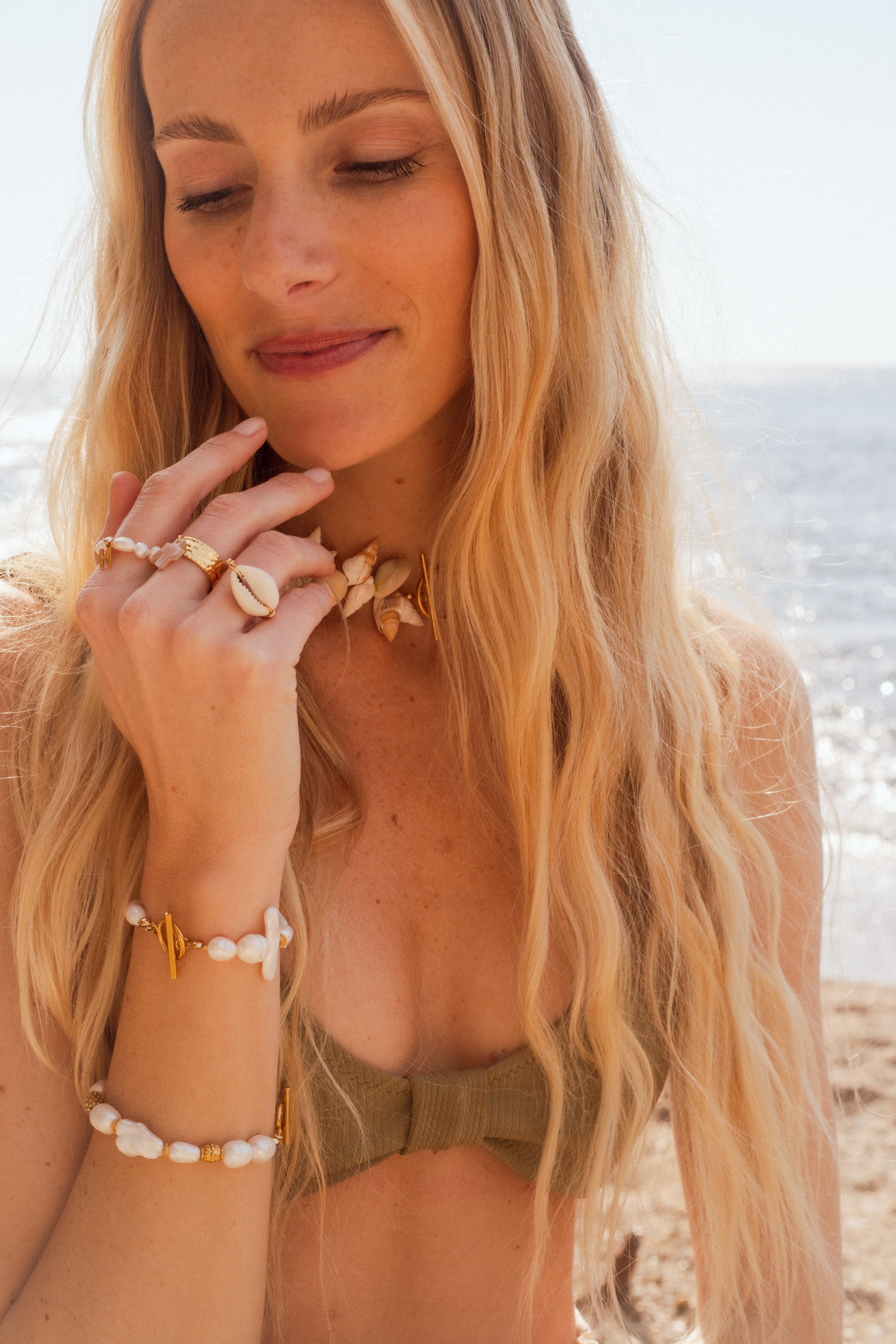 Portrait d'une femme aux cheveux longs et blonds, portant des bijoux en coquillage, sur une plage proche de la mer.