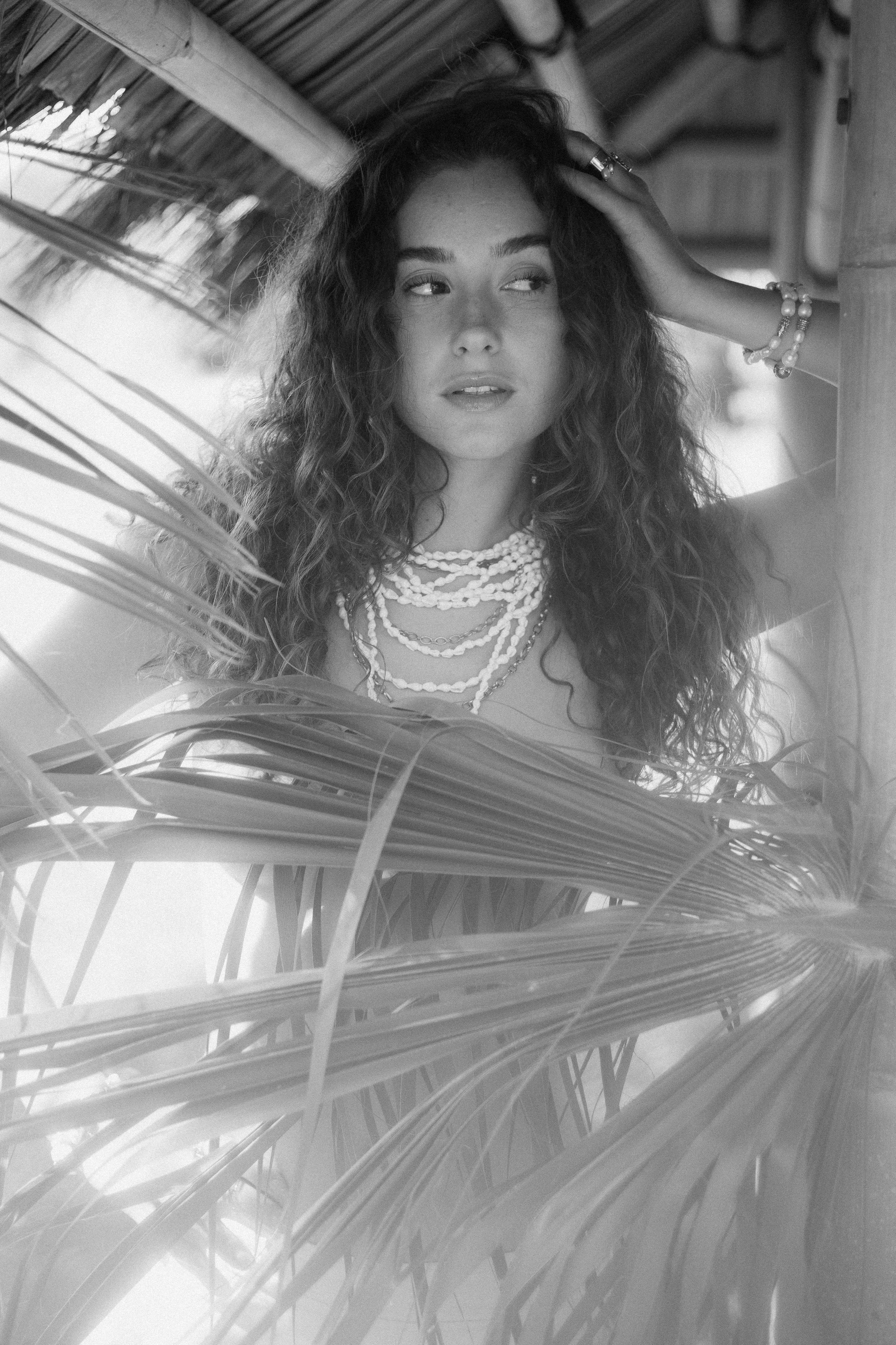 Une femme avec des cheveux bouclés portant plusieurs colliers et bracelets, se tenant dans un lieu en bois avec des éléments végétaux autour, vue en noir et blanc.