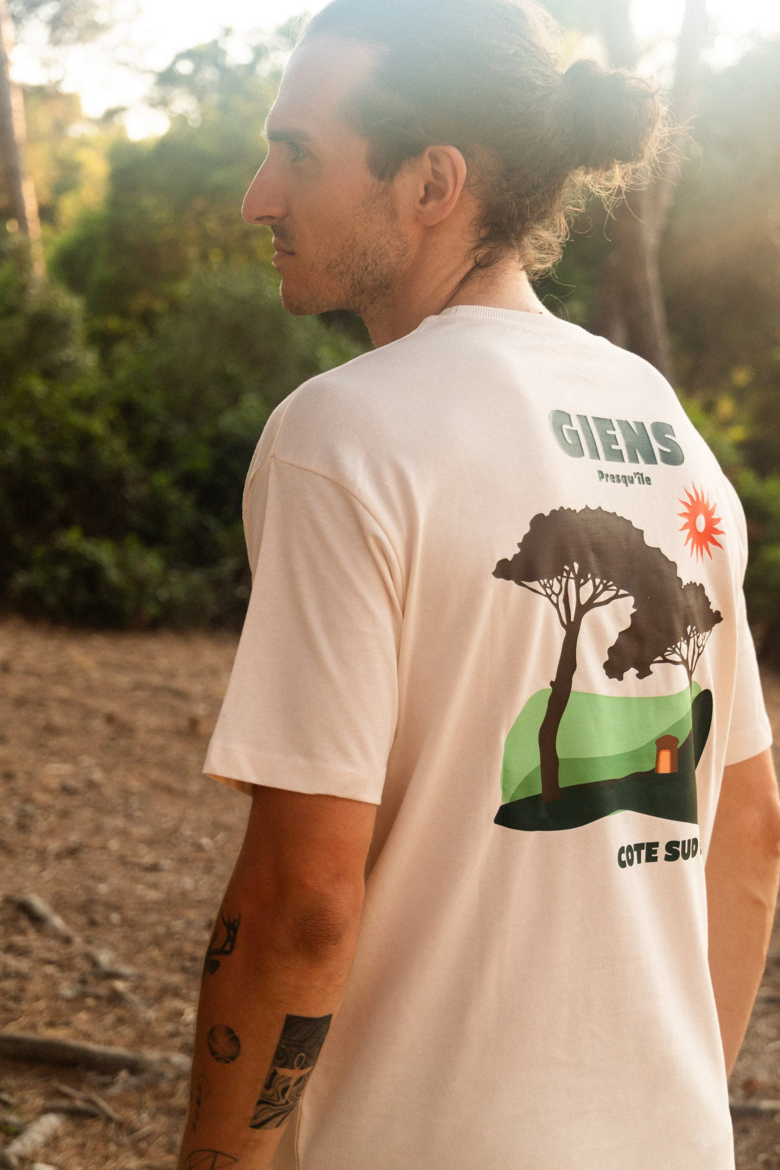 Un homme avec des tatouages sur le bras, vu de dos, portant un t-shirt beige avec un graphique représentant des arbres, un soleil rouge et un petit bâtiment au dos, en plein air dans la forêt pendant la journée.