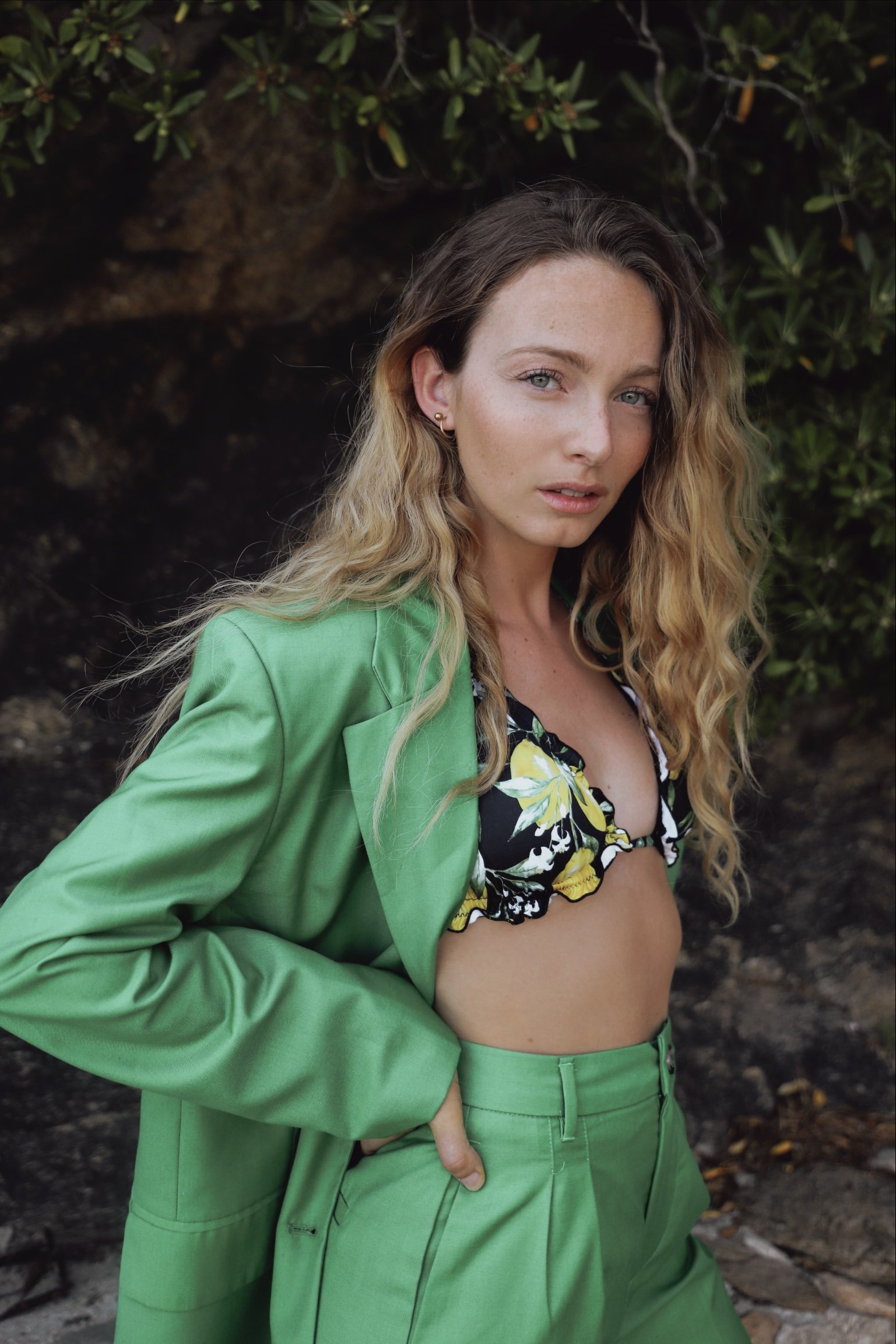 Jeune femme avec cheveux bouclés, portant un blazer vert, un bikini à motifs floraux noirs, blancs et jaunes, et un pantalon vert, posant devant un fond naturel avec des feuillages et des rochers.