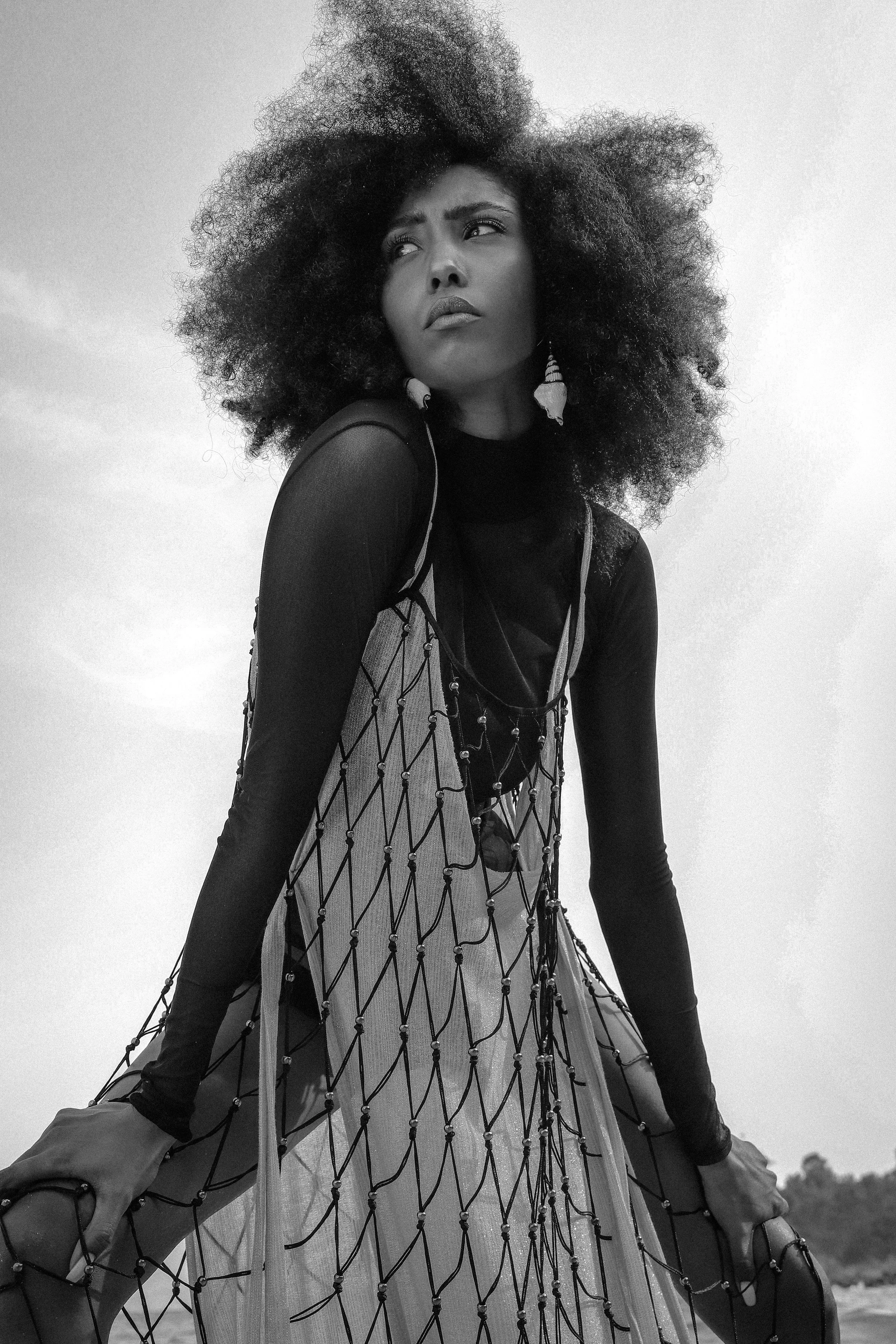Une femme noire avec des cheveux afro portant une robe avec un motif en filet, posant en extérieur, en noir et blanc.