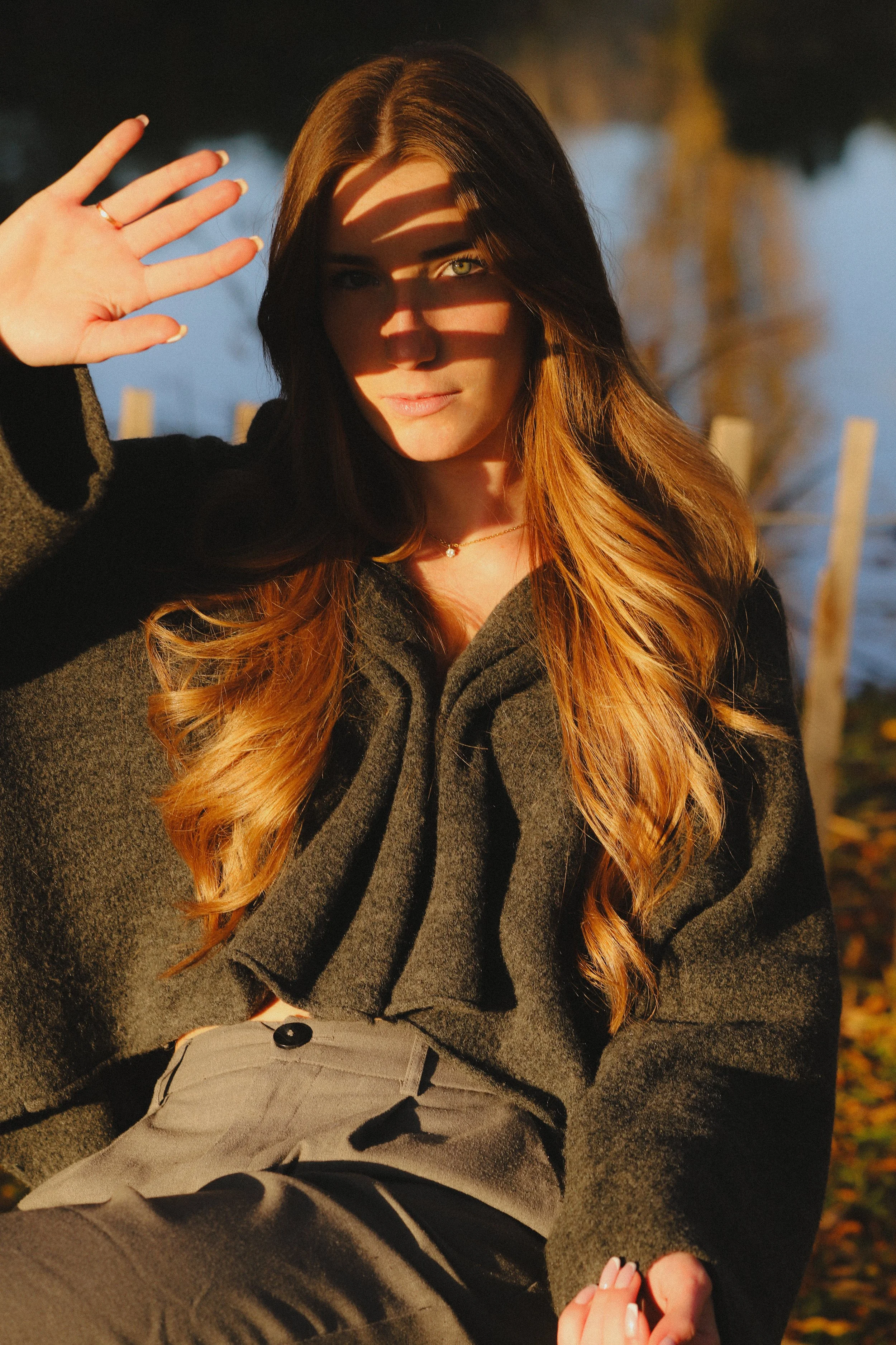 Jeune femme aux cheveux longs et bouclés, portant un manteau gris foncé, assise à l'extérieur avec un lac ou une rivière en arrière-plan, faisant un geste de salut avec la main gauche, dans la lumière dorée du coucher de soleil.