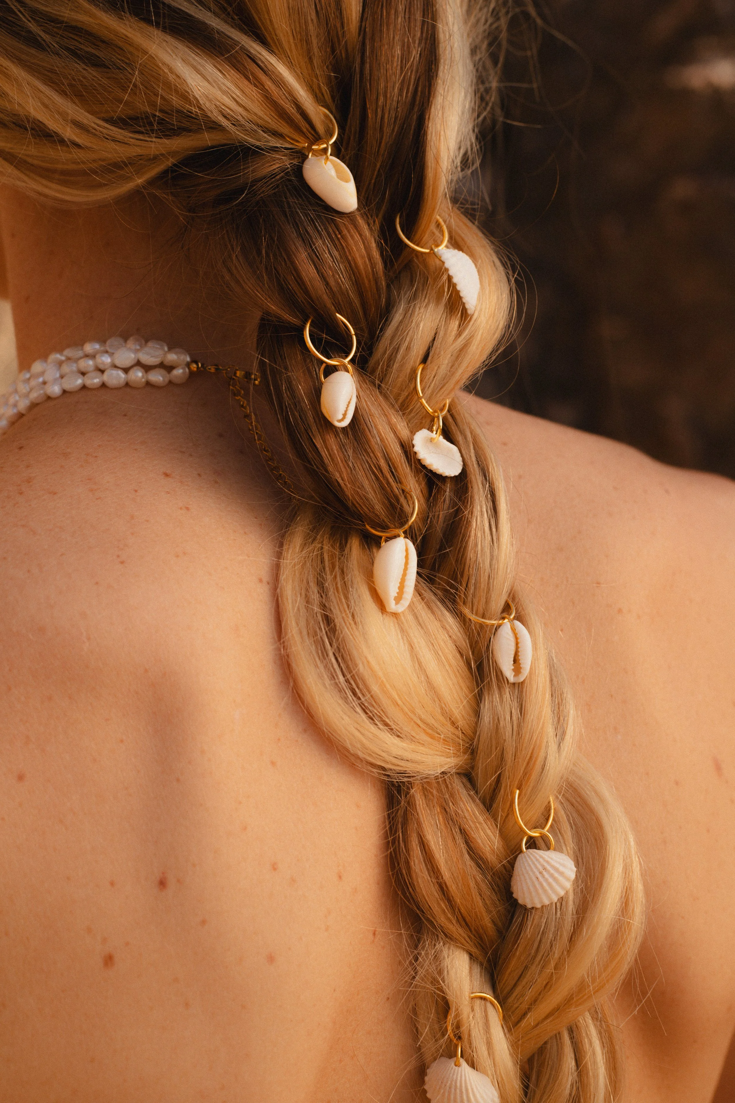 Coiffure en tresse de cheveux blonds décorée avec des coquillages blancs et des anneaux dorés, portant un collier de perles autour du cou.