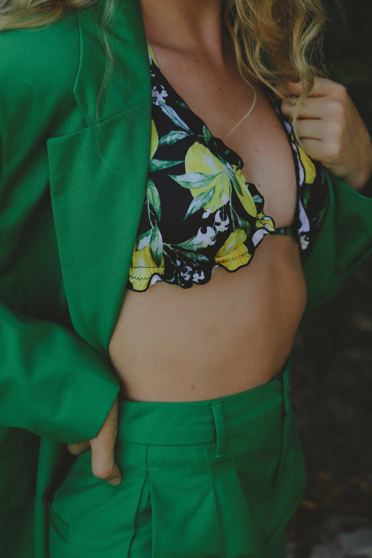 Une femme portant un blazer vert, un haut à fleurs coloré et un pantalon vert, avec une main dans la poche du pantalon.