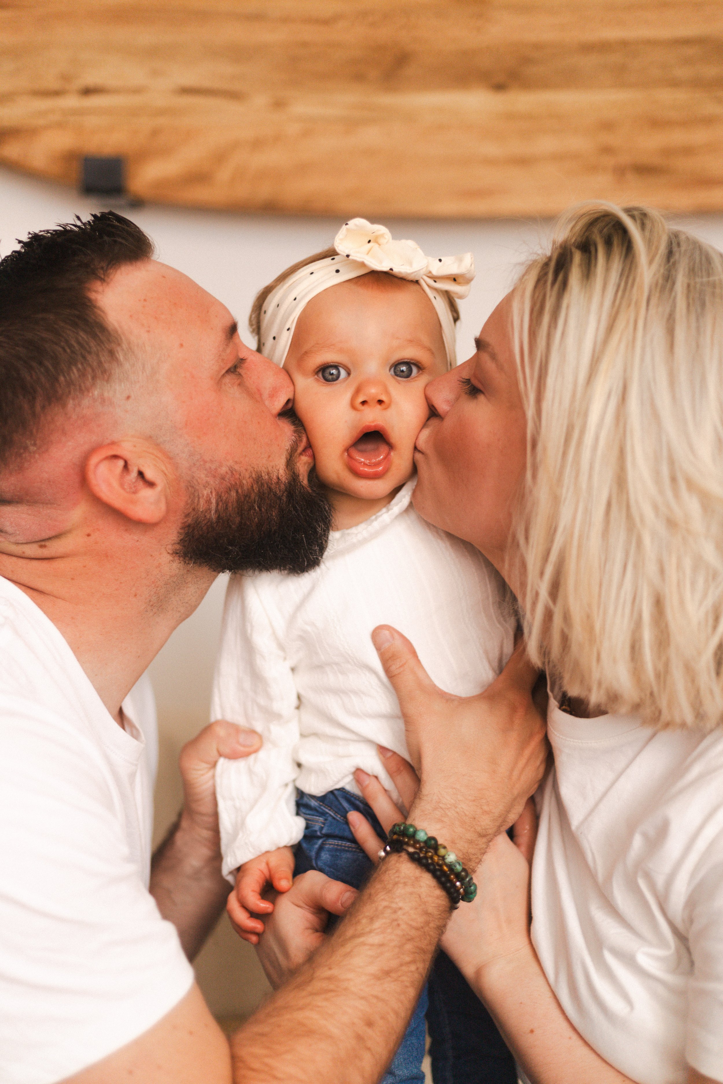 Un bébé entre un homme et une femme, tous deux l'embrassant sur les joues. La bébé a les yeux grands ouverts et porte un bandeau blanc avec des points noirs.
