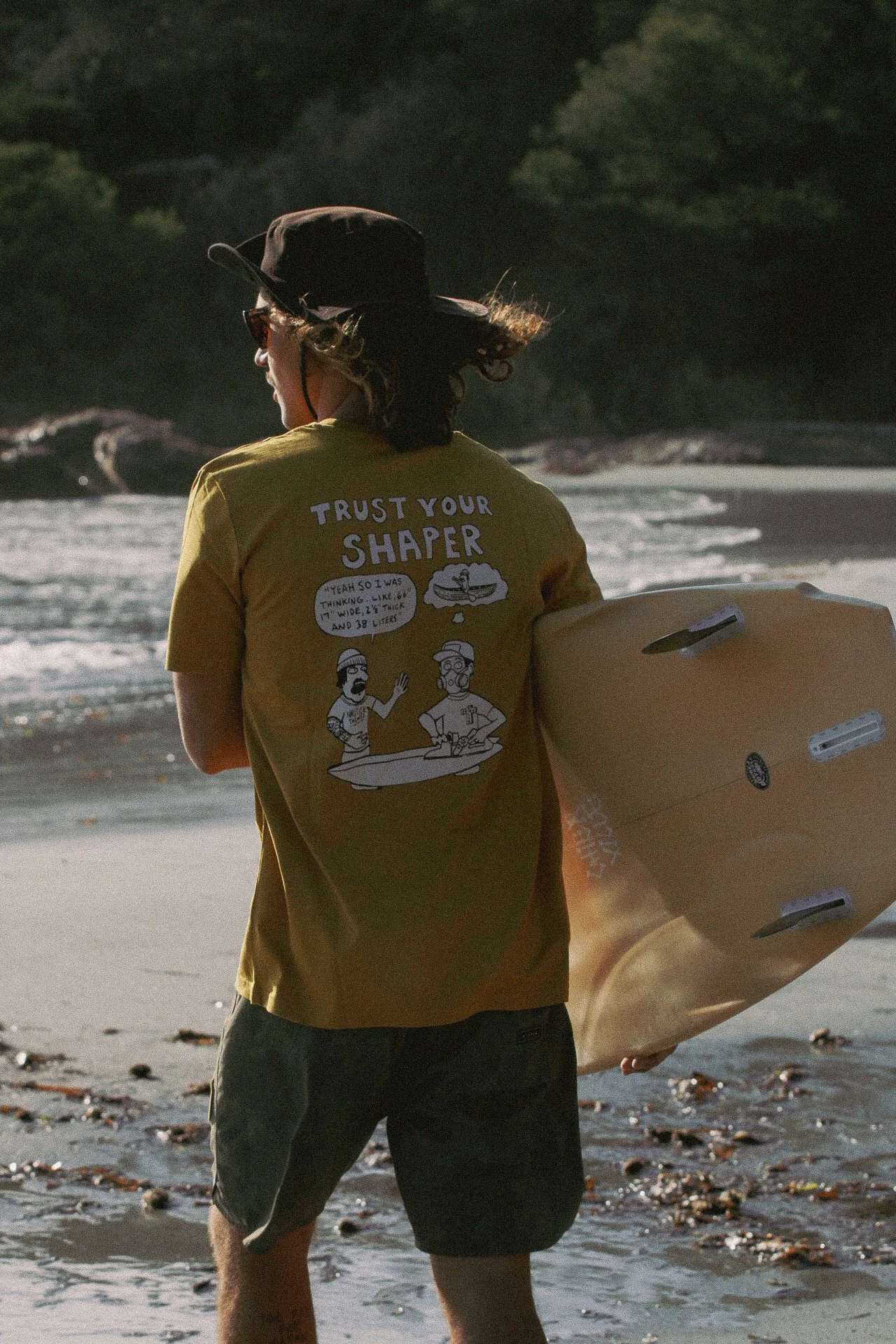 Une personne de dos portant un t-shirt jaune avec une illustration de deux personnes discutant, tenant une planche de surf à la plage, avec des vagues et des arbres en arrière-plan.