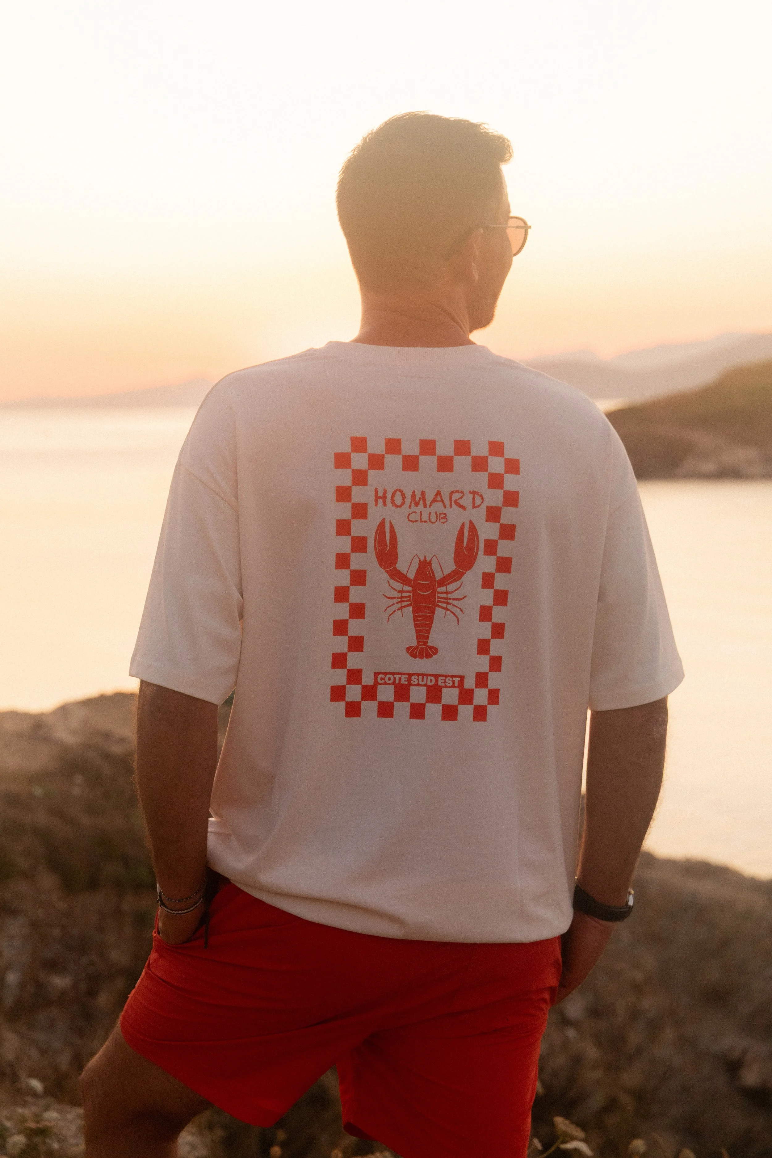 Un homme debout sur une côte rocheuse lors d'un coucher de soleil, vu de dos, portant un t-shirt blanc avec un crabe rouge et un motif à carreaux. Le t-shirt dit "HOMARD CLUB COTE SUD EST".