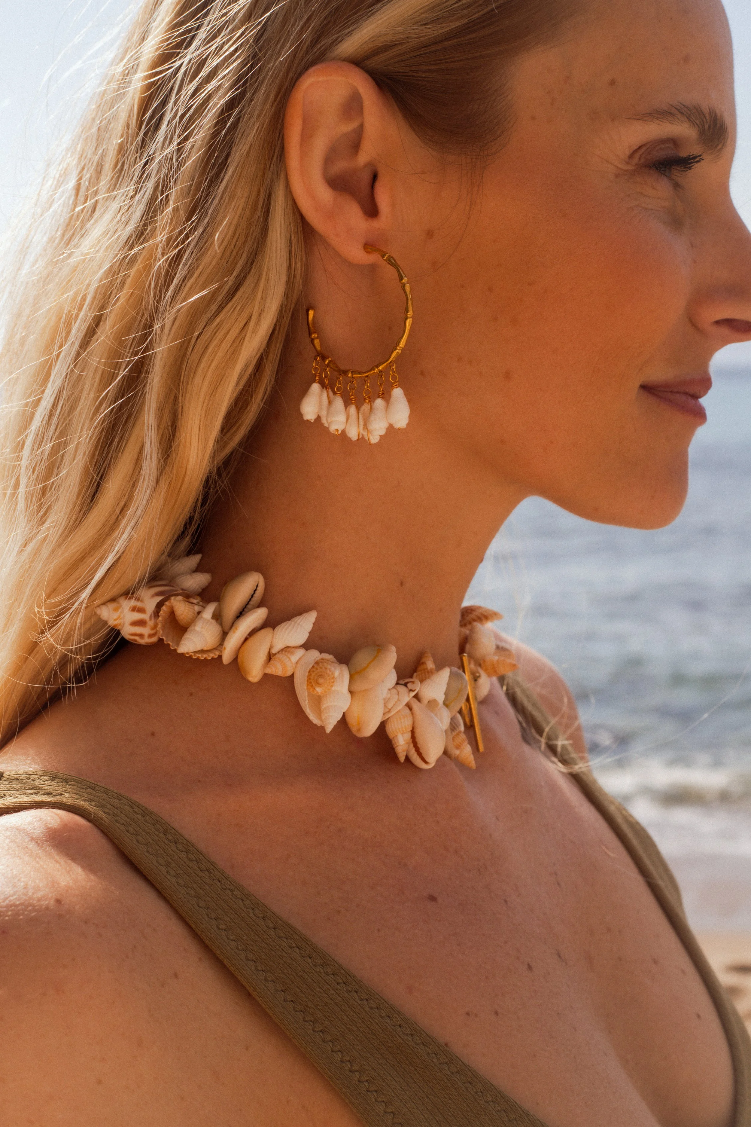 Une femme souriante portant un collier de coquillages et des boucles d'oreilles avec des coquillages au bord de la mer.
