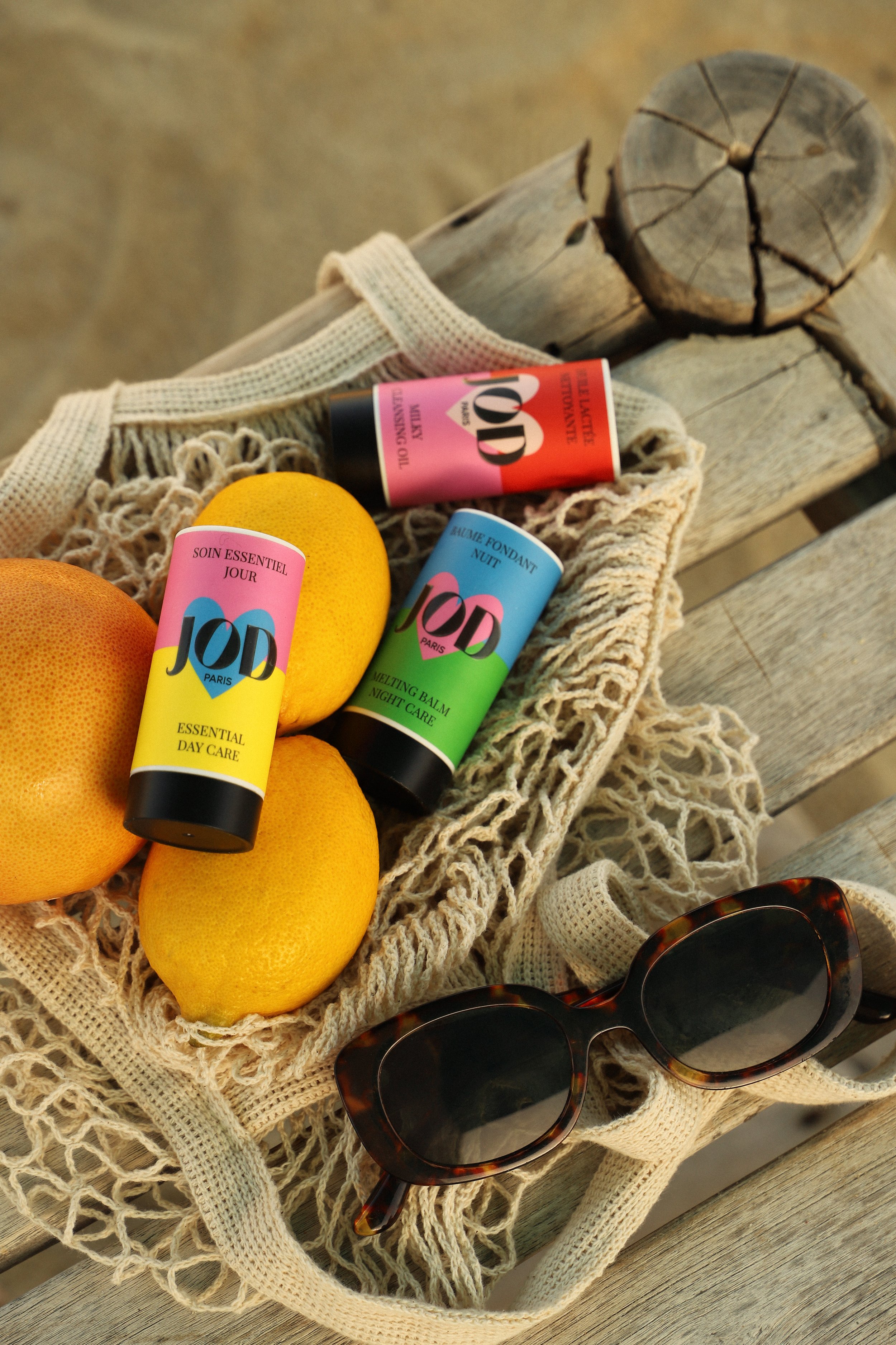 Une image montre un panier en coton contenant trois citrons, deux tubes de produits de soin JOD en couleur rose et jaune, ainsi qu'une paire de lunettes de soleil à monture écaille, le tout posé sur un banc en bois.