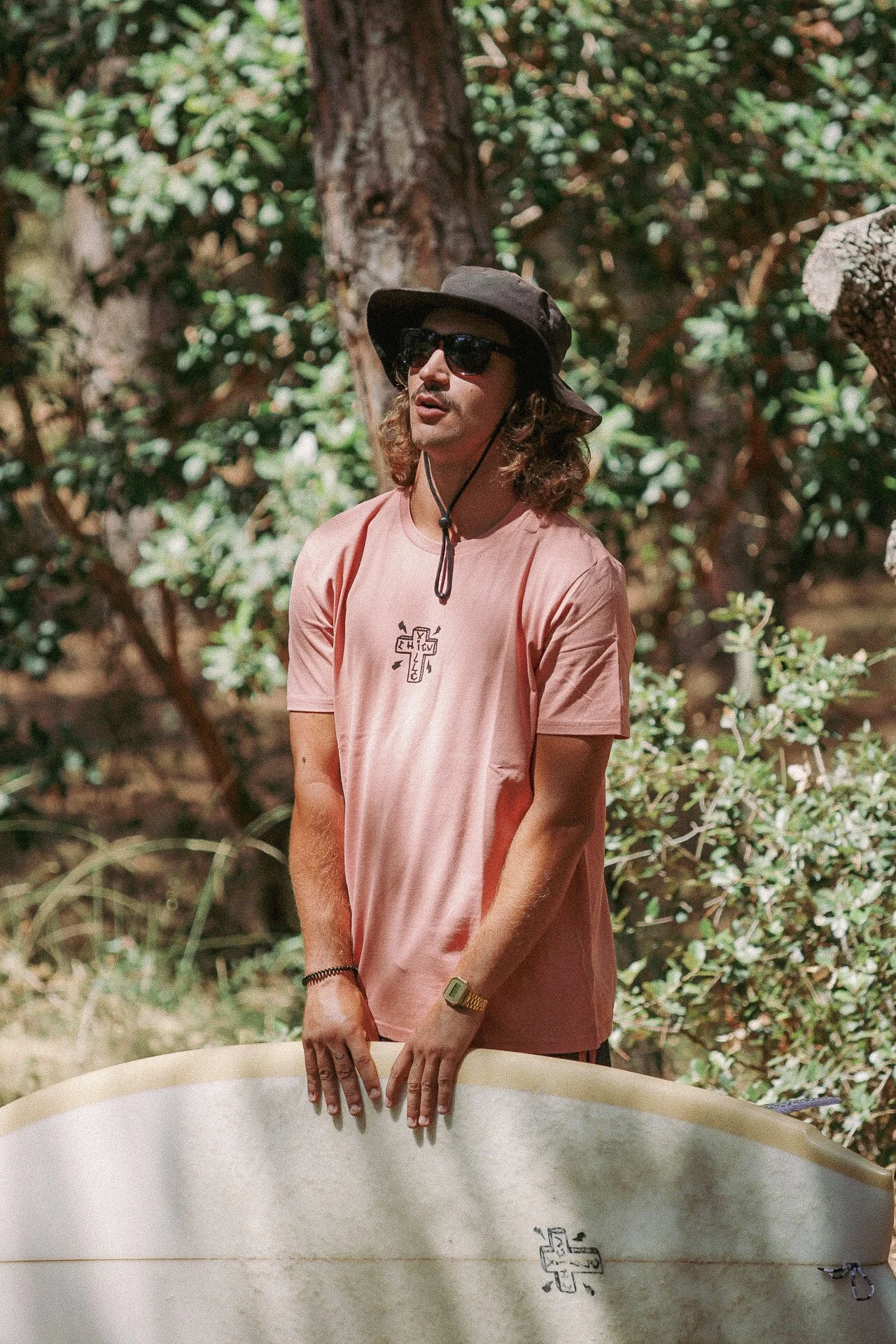 Jeune homme avec chapeau, lunettes de soleil et t-shirt rose pâle, tenant une planche de surf, dans un environnement naturel avec arbres et buissons.