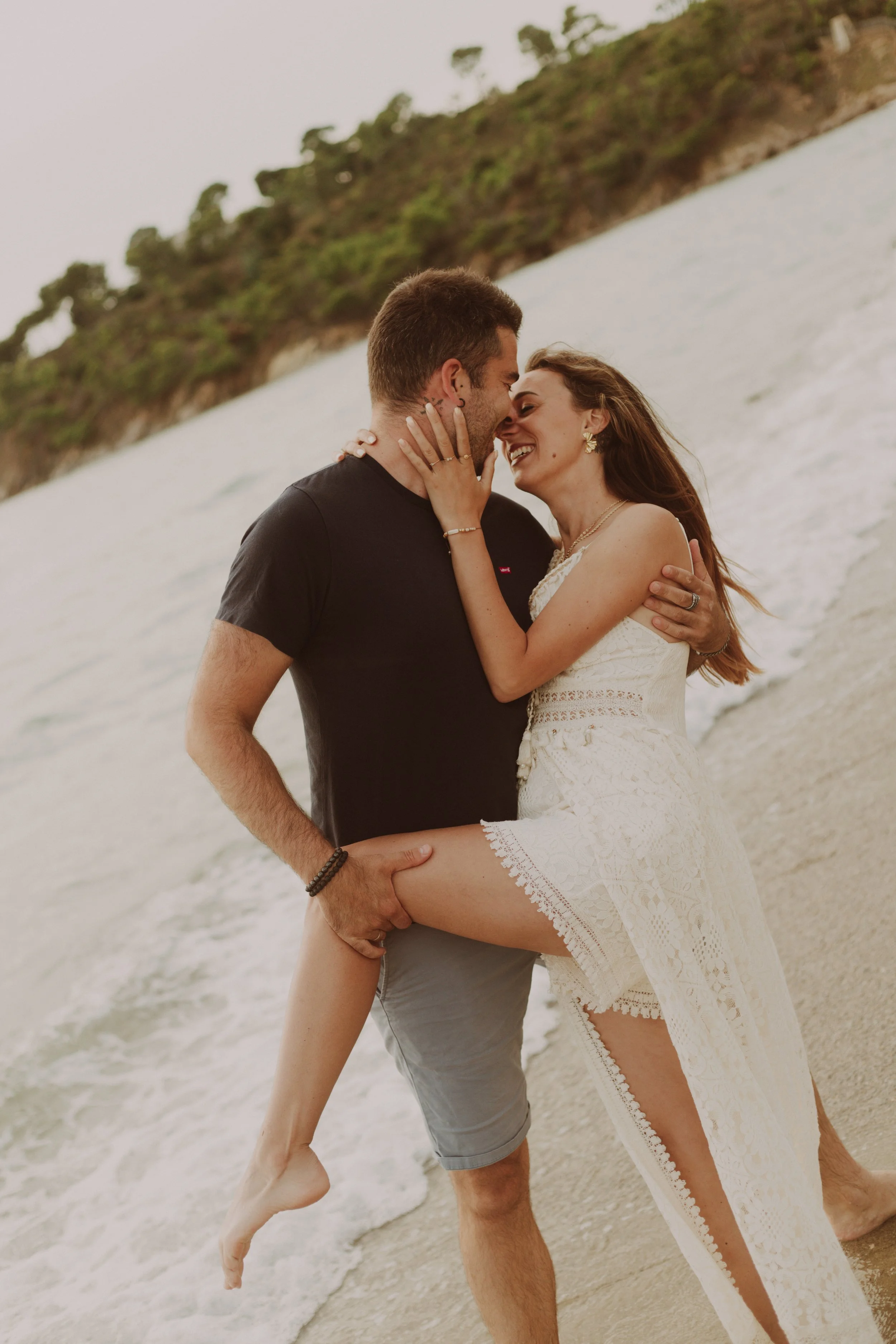 Un couple en tenue estivale partage un moment romantique sur la plage au coucher du soleil, avec la mer en arrière-plan.