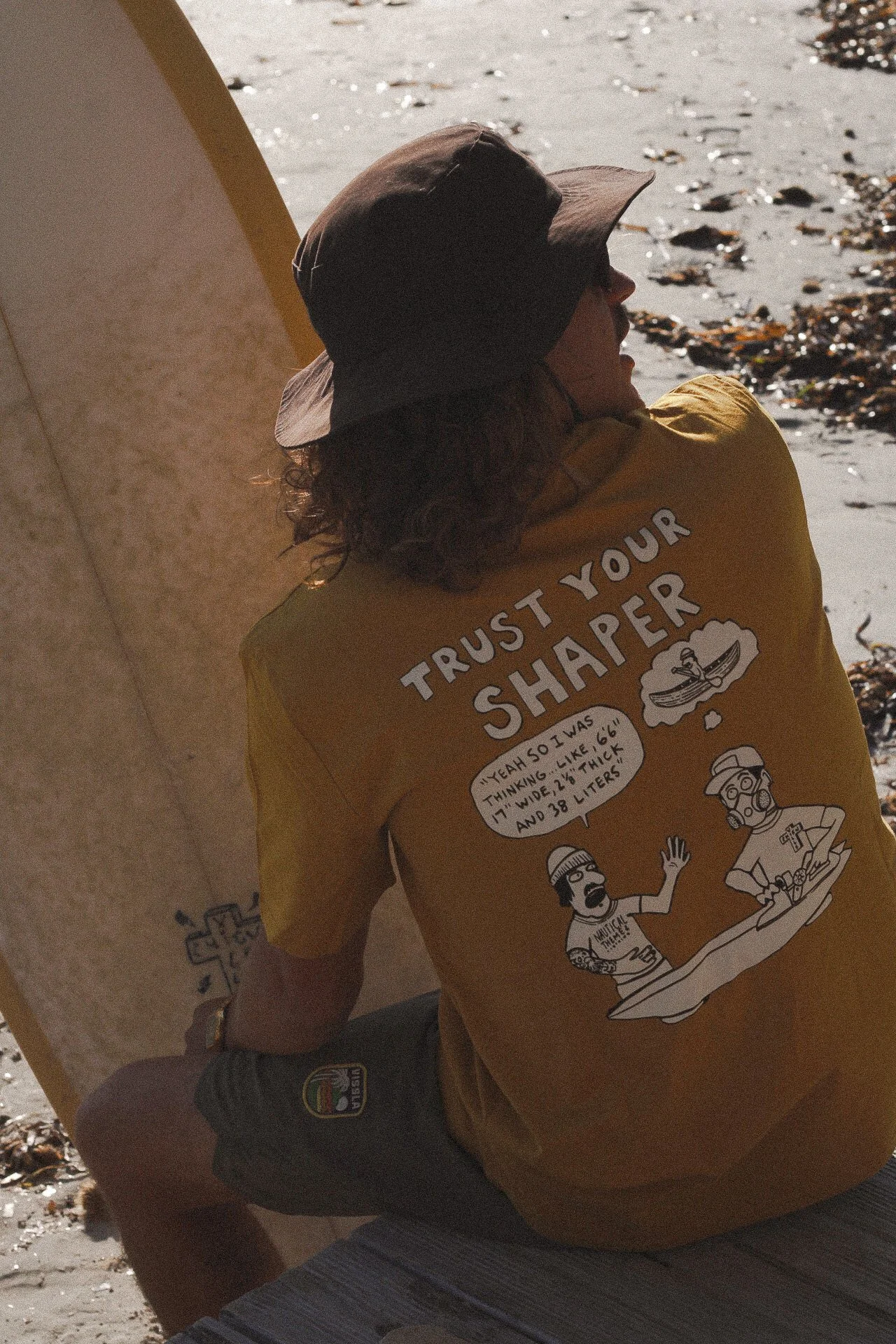 Une personne assise sur une plage, vue de dos, portant un t-shirt jaune avec des illustrations et du texte, avec des vagues et des algues en arrière-plan.