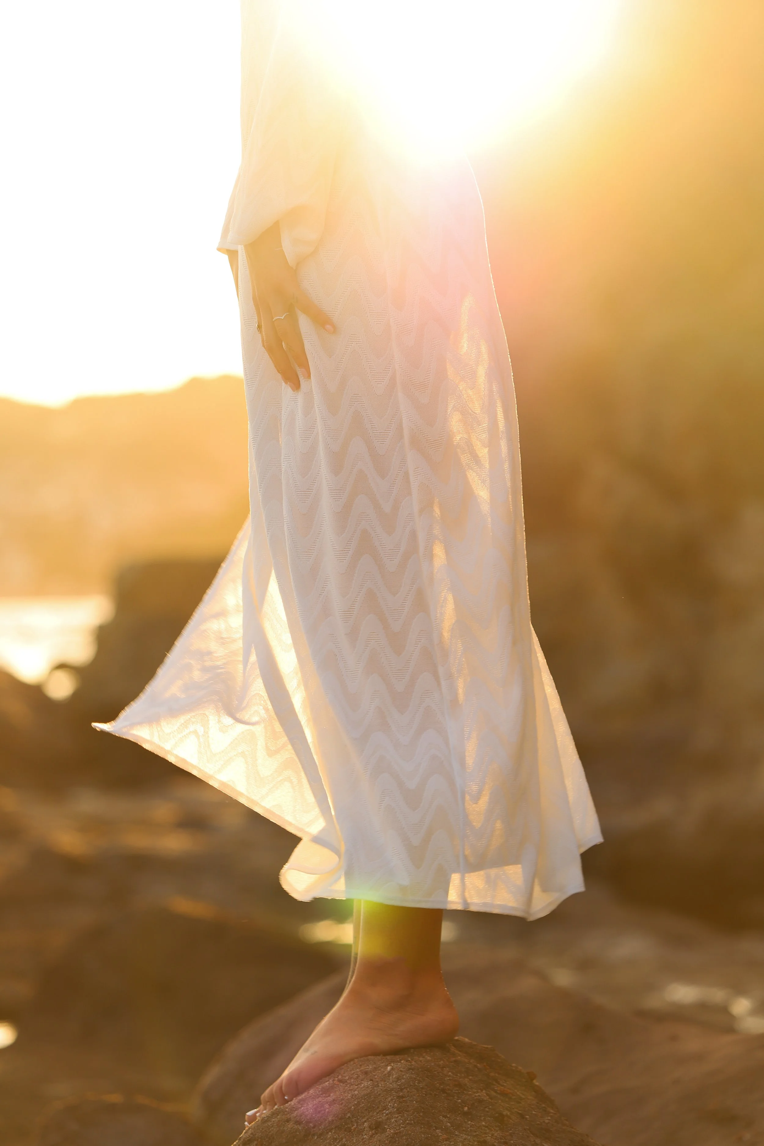Une personne debout sur un rocher, portant une robe blanche translucide, au coucher du soleil.