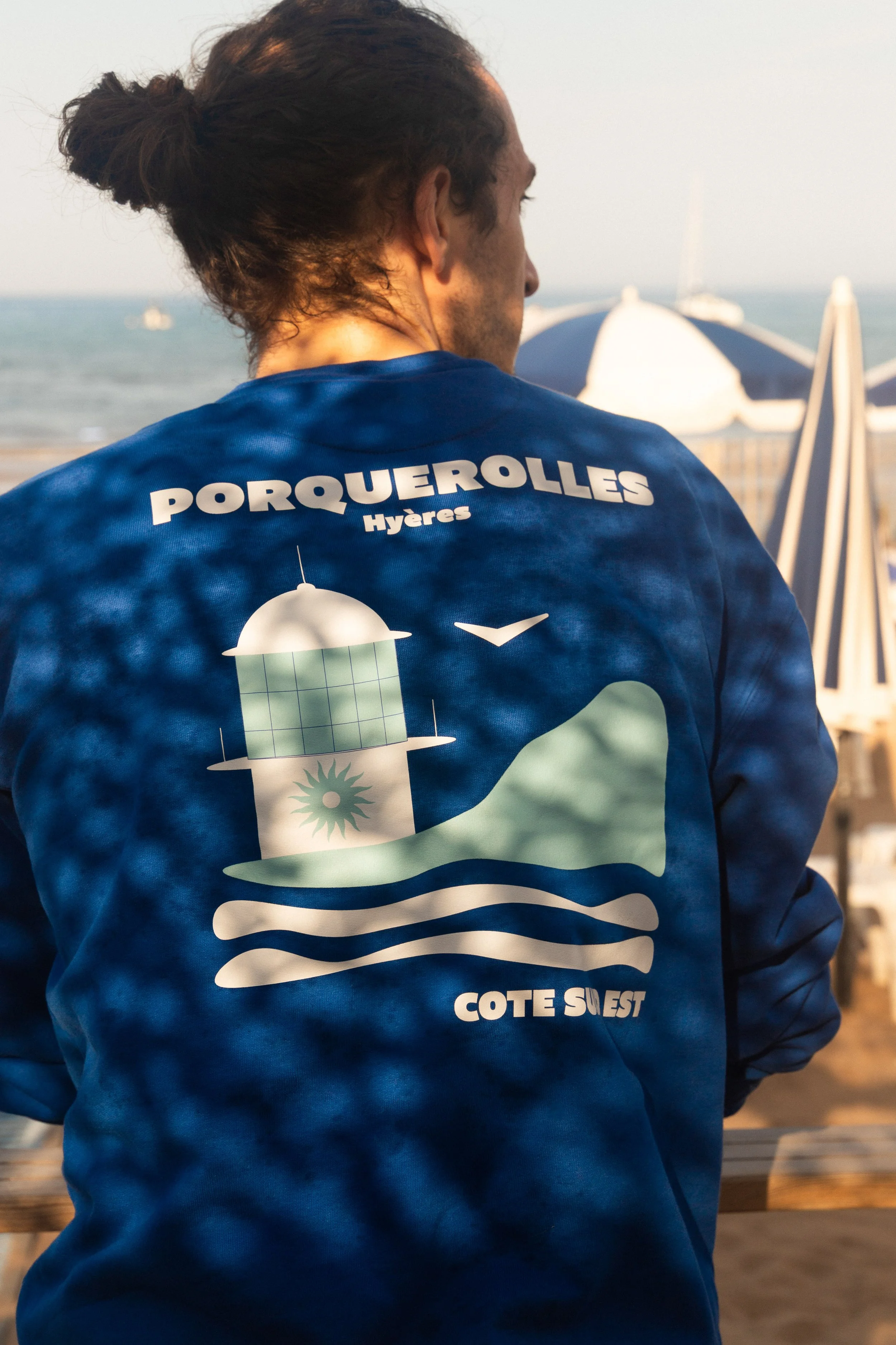 Homme vu de dos portant un sweat-shirt bleu avec un motif représentant un phare, une mer et un oiseau, sur la côte sud-ouest de Porquerolles, en Méditerranée.