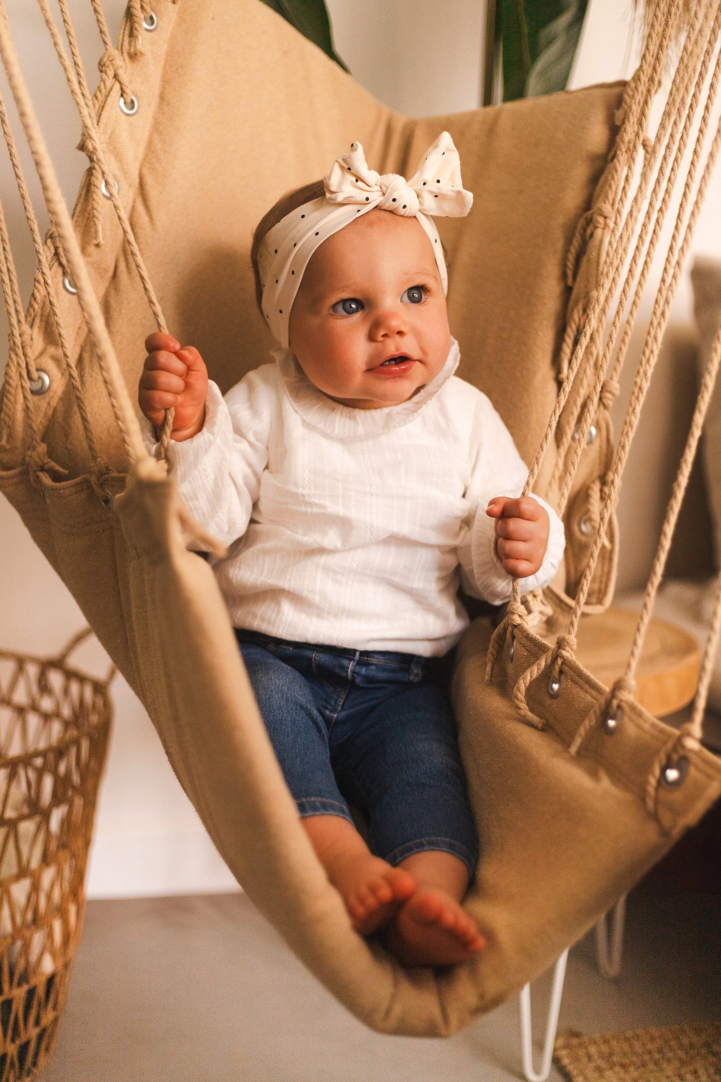 Un bébé assis dans une balançoire suspendue en tissu beige, portant un bandeau blanc avec des points noirs, une blouse blanche et un pantalon en jean, dans un intérieur chaleureux.