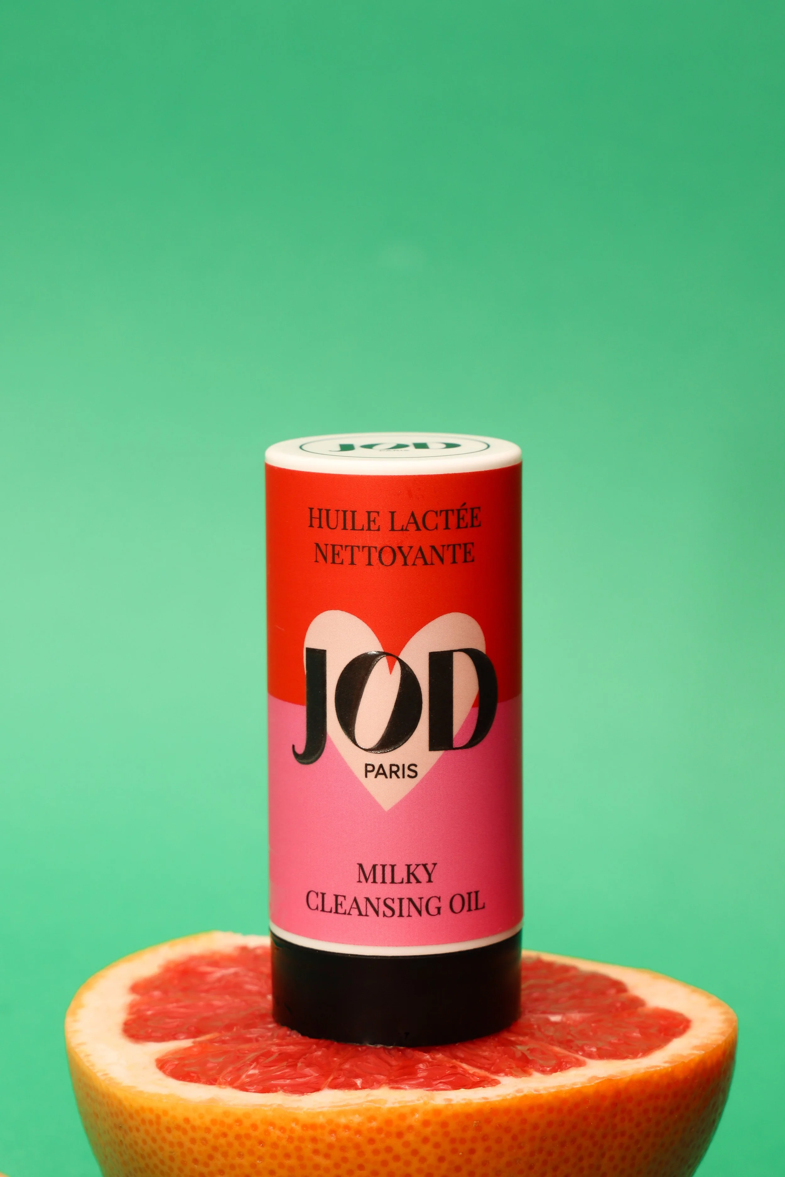 Un produit de nettoyage à l'huile lactée JOD Paris, placé sur une moitié de pamplemousse, avec un fond vert dégradé.