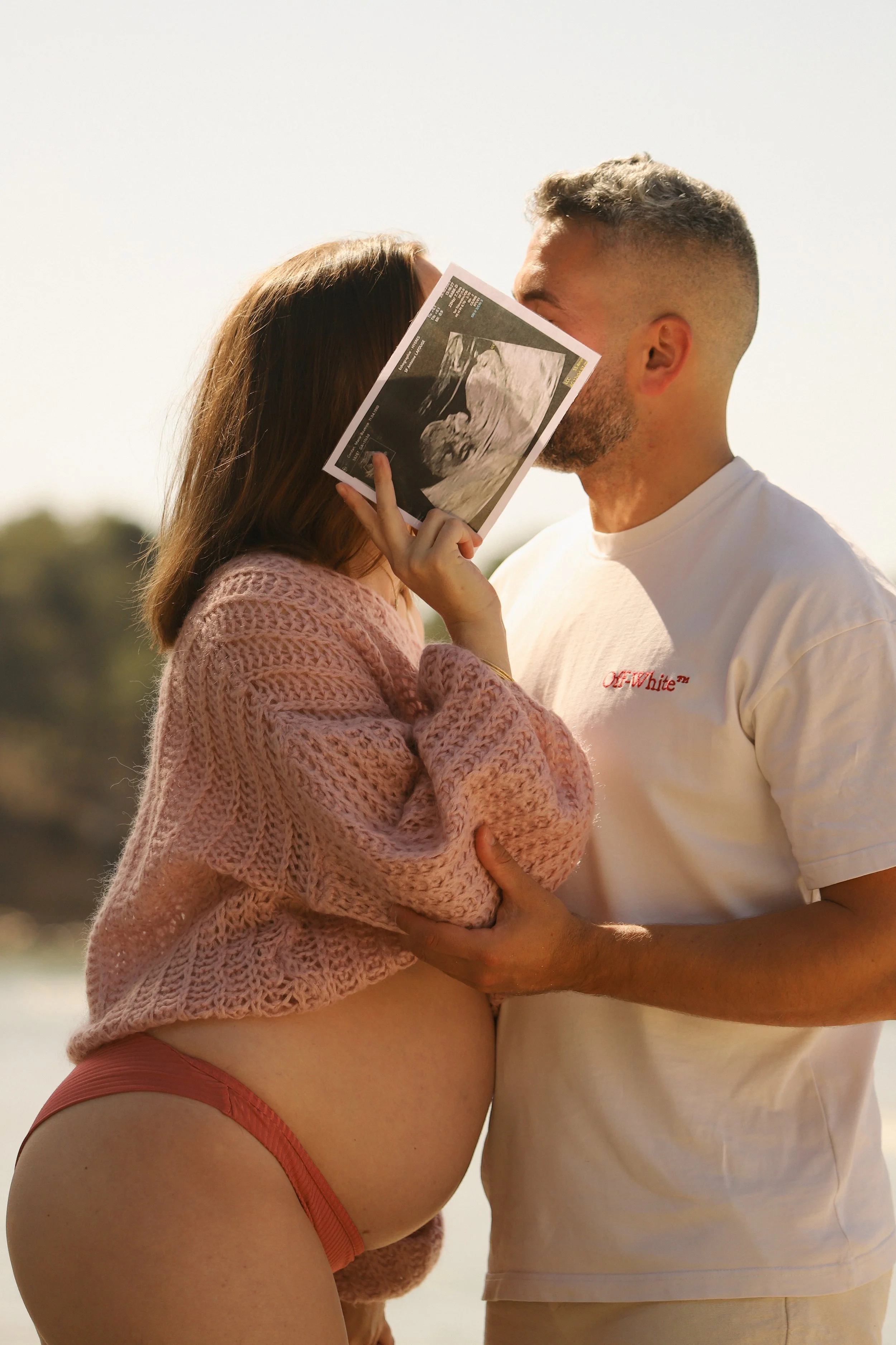 Un couple attend un bébé, la femme tenant une échographie devant le visage du partenaire, lors d'une séance photo en extérieur.