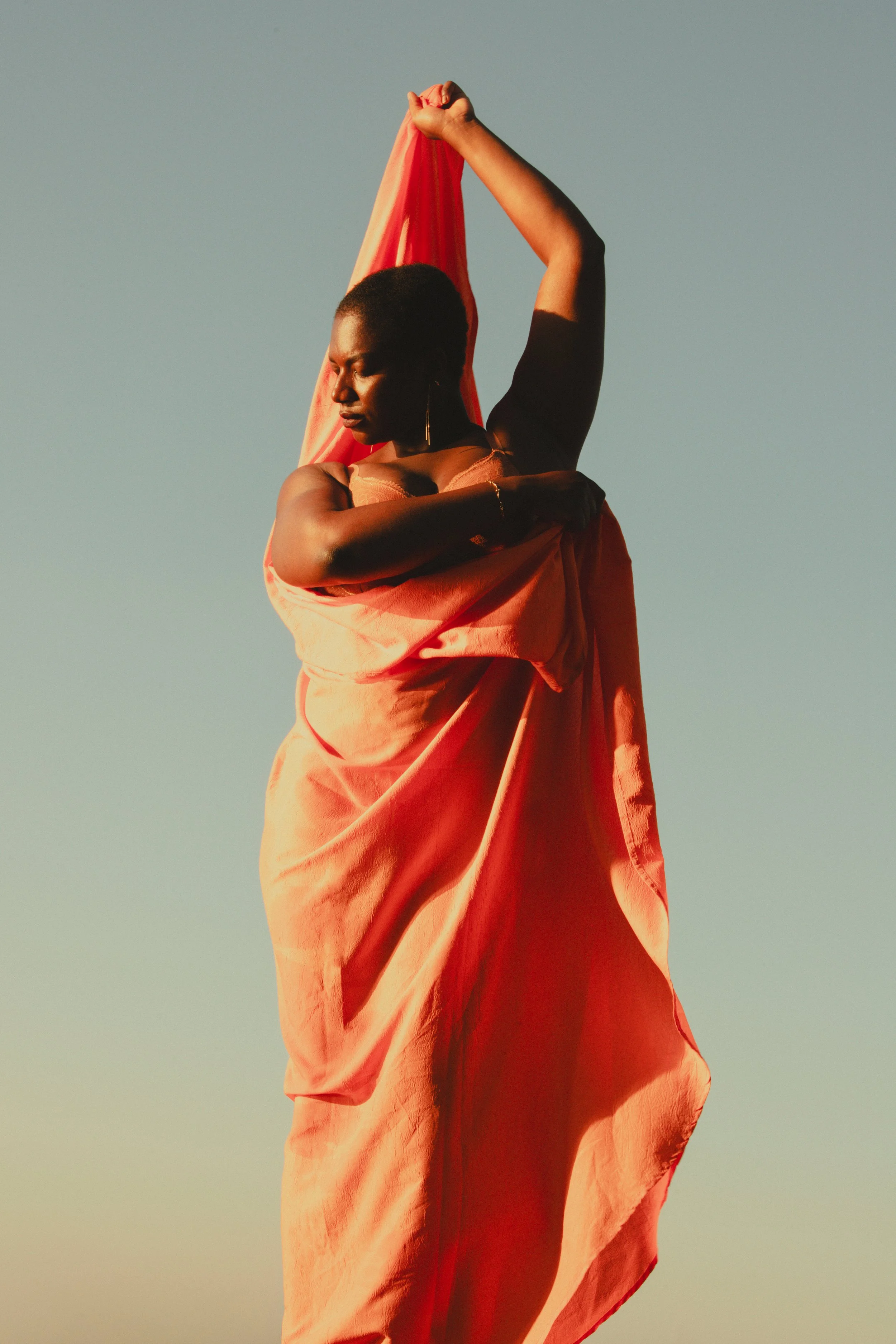 Femme noire dans une robe orange, levant un bras avec un tissu, au coucher du soleil.