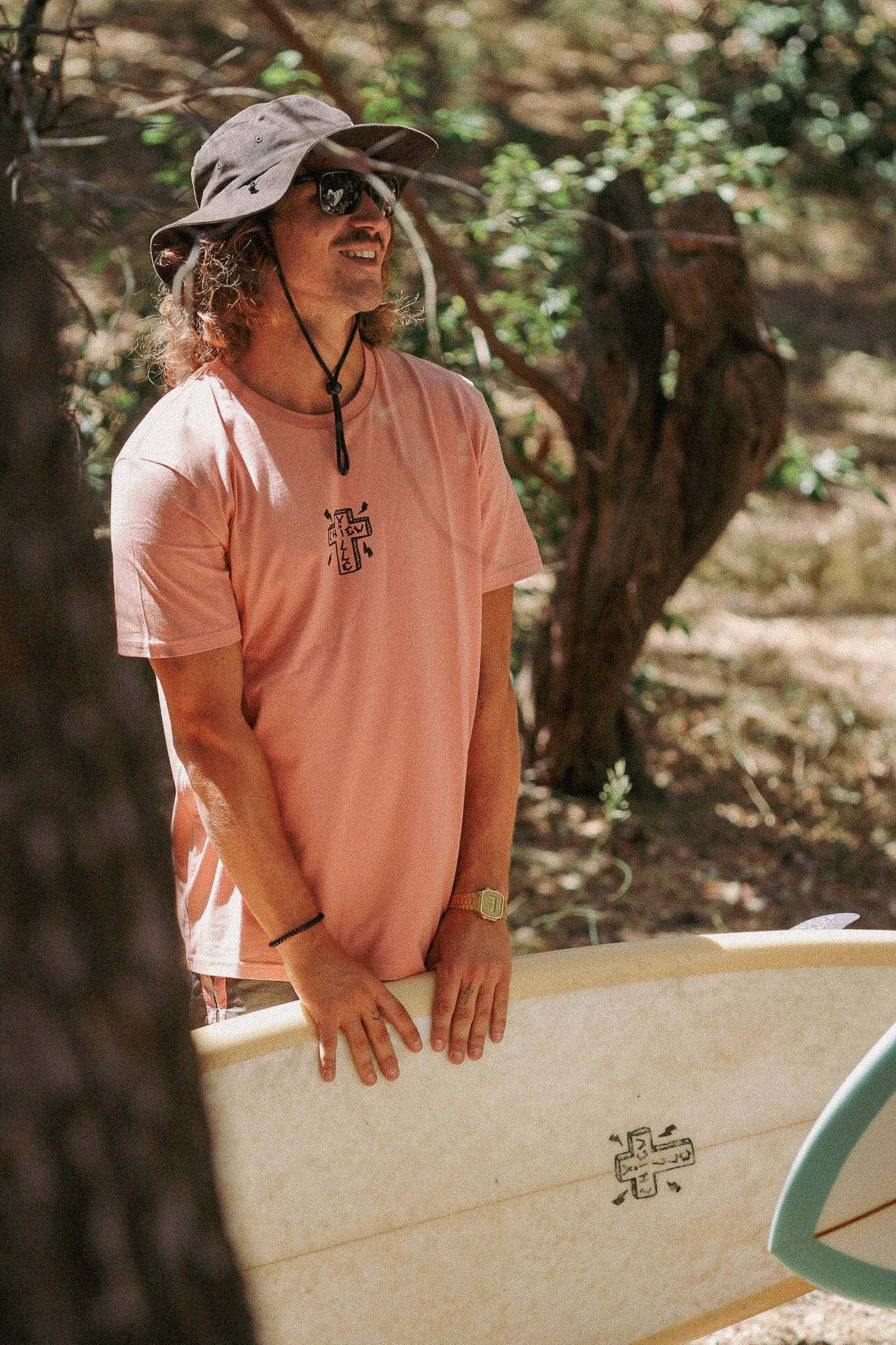 Une personne portant un chapeau à large bord, des lunettes de soleil, une chemise rose clair, tient une planche de surf dans un environnement boisé.